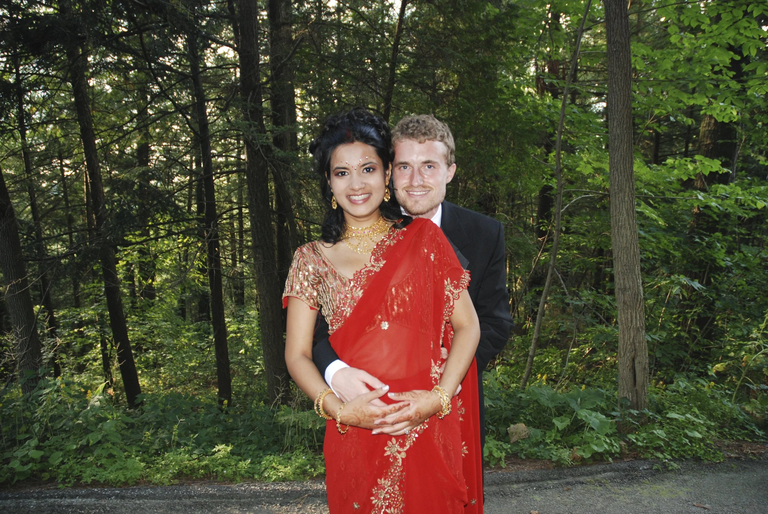 A couple stands together outdoors in a forest, dressed in formal attire. The woman wears a red and gold sari with jewelry, and the man wears a black tuxedo. They are smiling, with the man's arms around the woman. Kleinburg Ontario.