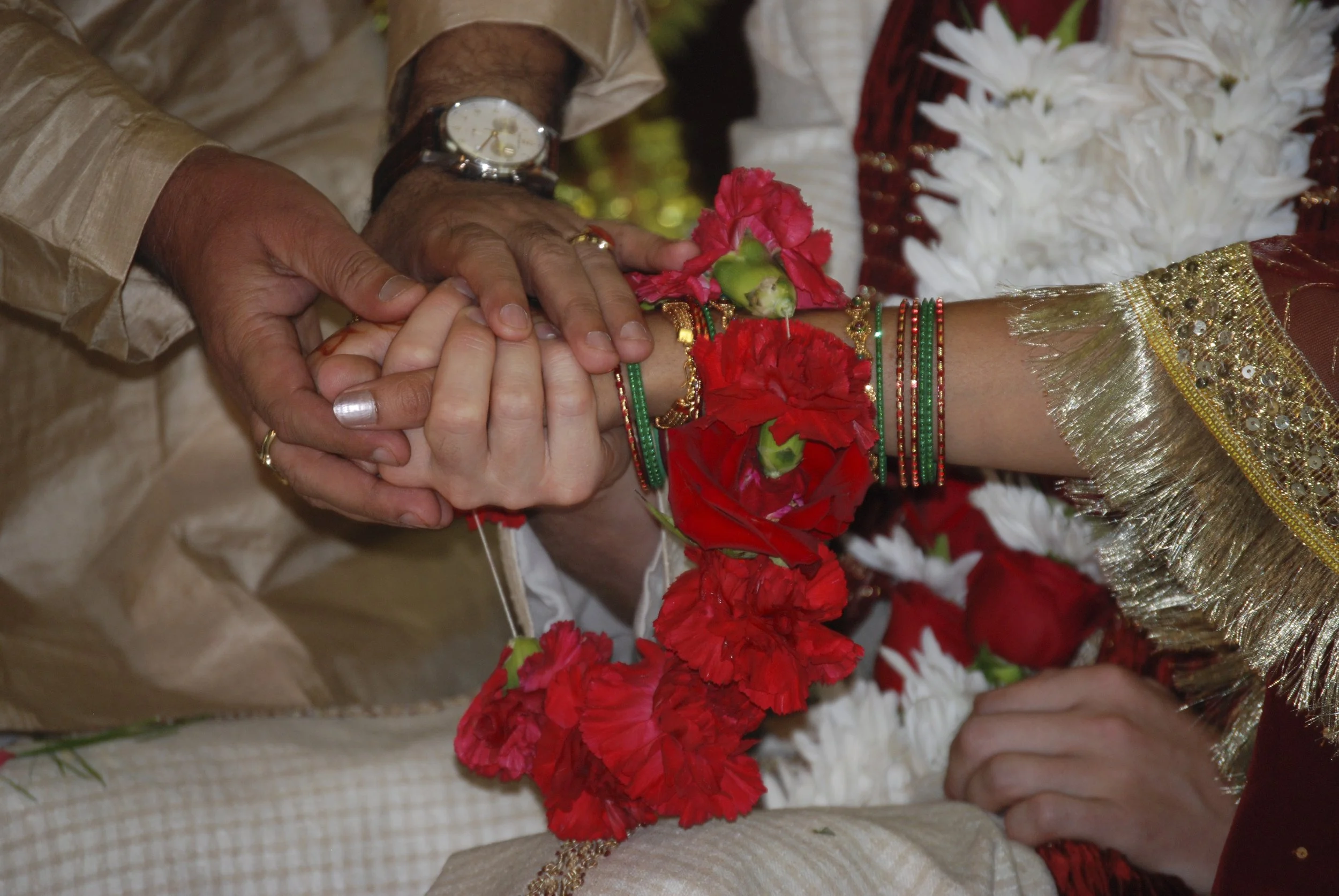Close-up of a traditional Hindu wedding ceremony where the bride and groom are holding hands, adorned with colourful bangles, jewelry, and red flowers, with a background of white flowers and decorative attire Oakville Ontario.