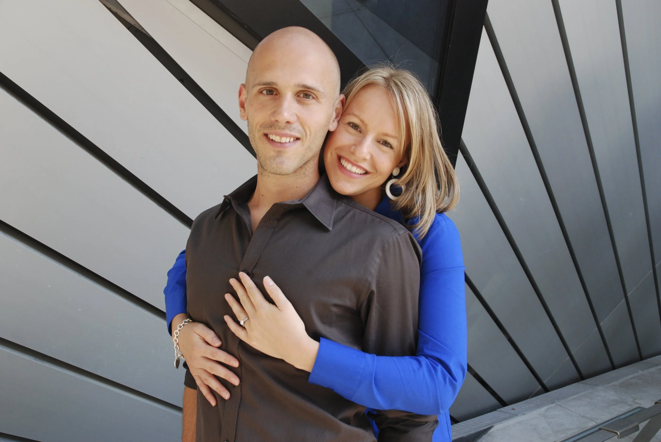 A man and woman smiling with the woman hugging the man from behind, standing in front of a modern building.