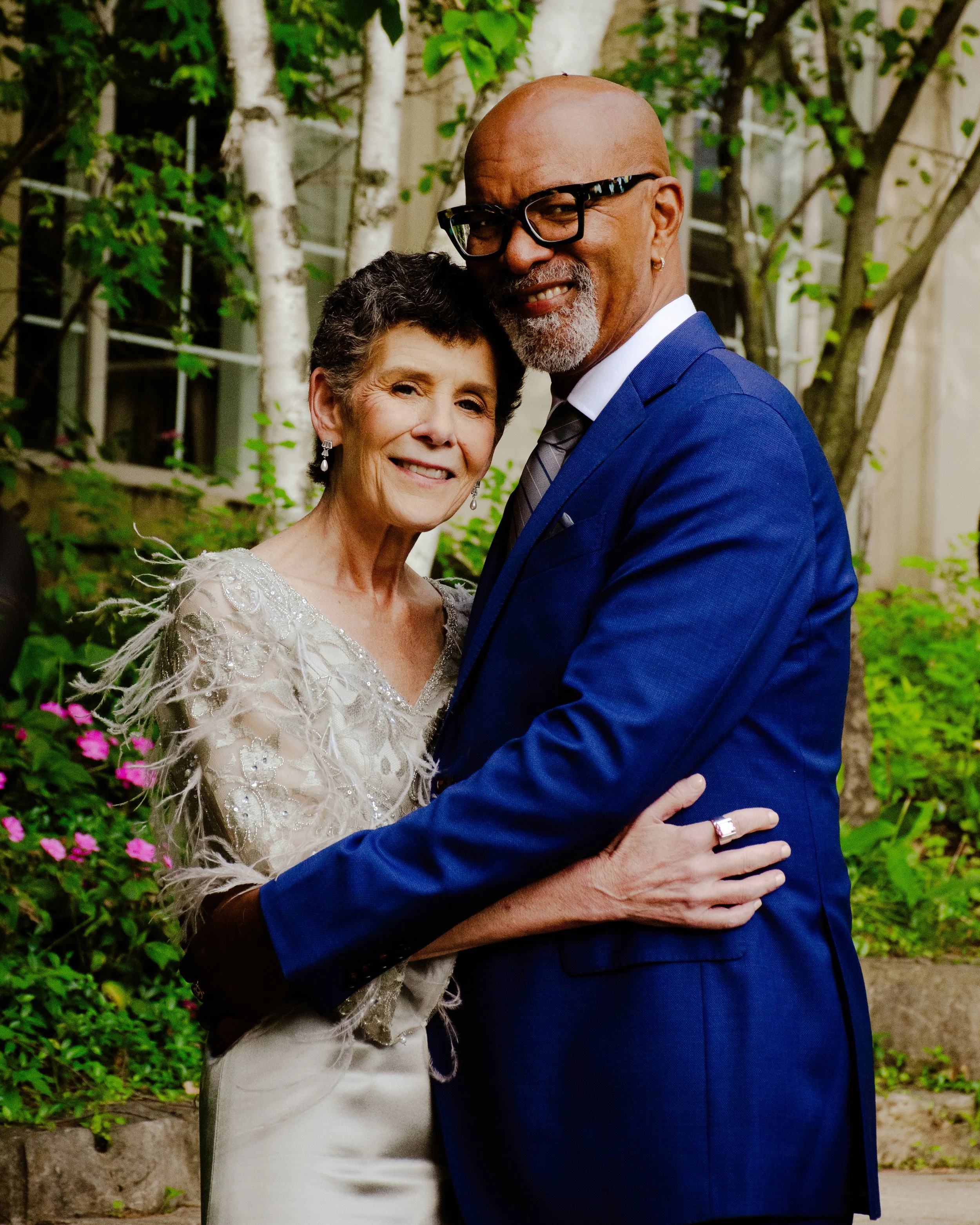An older senior bride and an older groom embracing outdoors, both smiling. The white woman  is wearing a cream-coloured wedding dress with intricate embroidery and feather details. The black man is bald, wearing glasses, a blue suit, white shirt,
