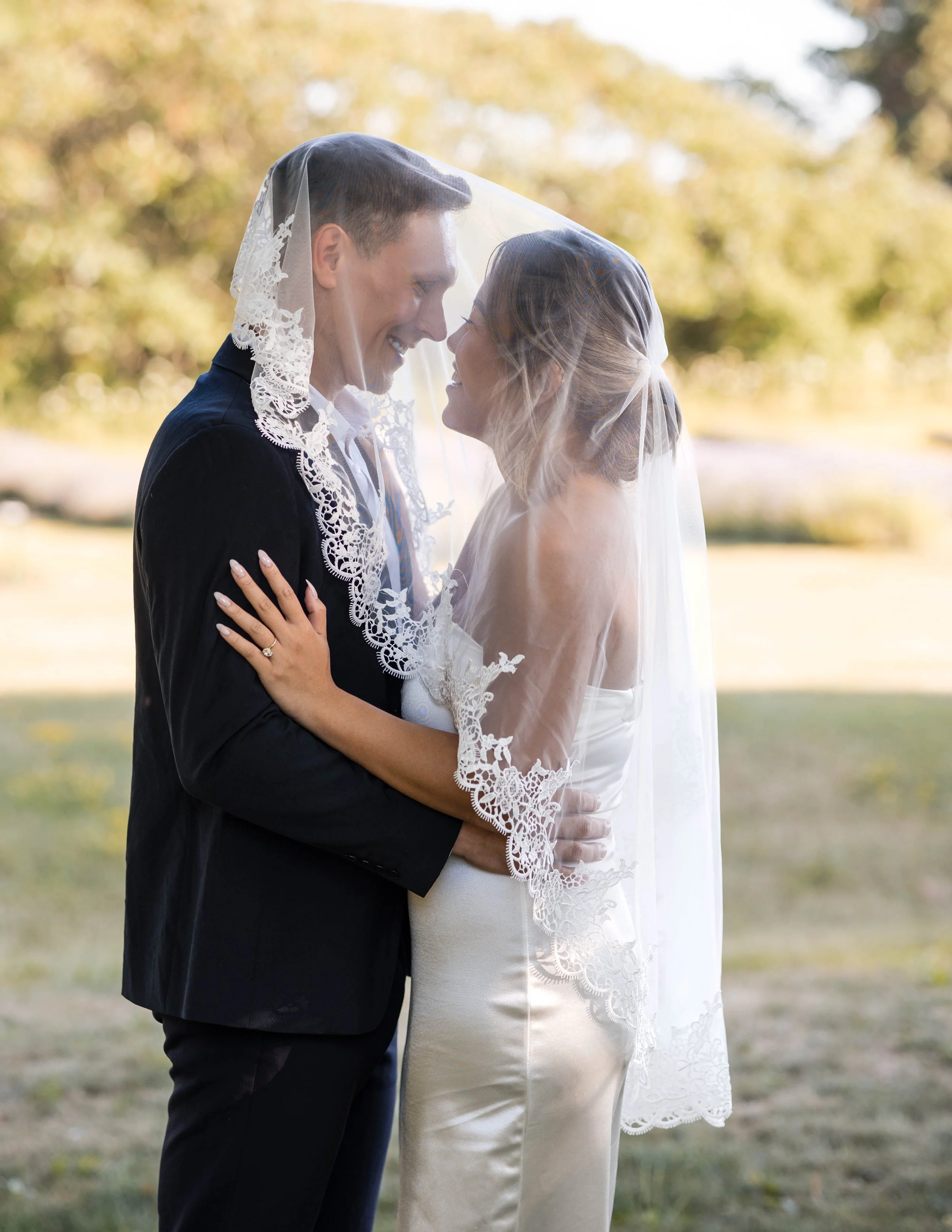 A bride and groom are close together outdoors, sharing a kiss under a lace wedding veil during sunset, with trees and grass in the background at Tranquility Trails Farm in Pickering Ontario.