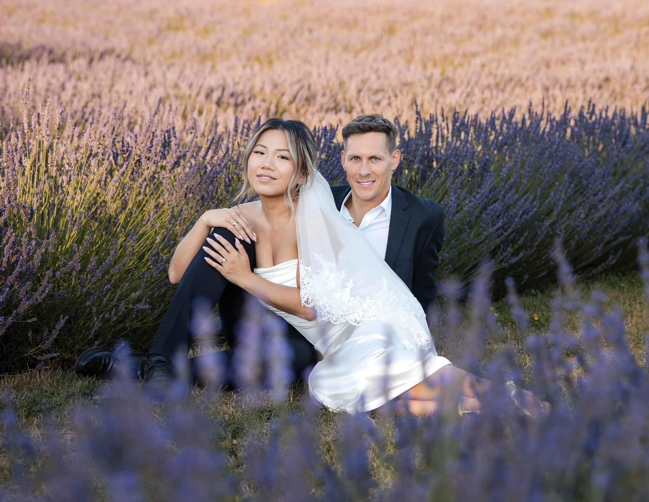A newlywed couple sitting on the grass in a lavender field during sunset, smiling and posing for a wedding photo at Tranquility Trails Farm in Pickering Ontario.