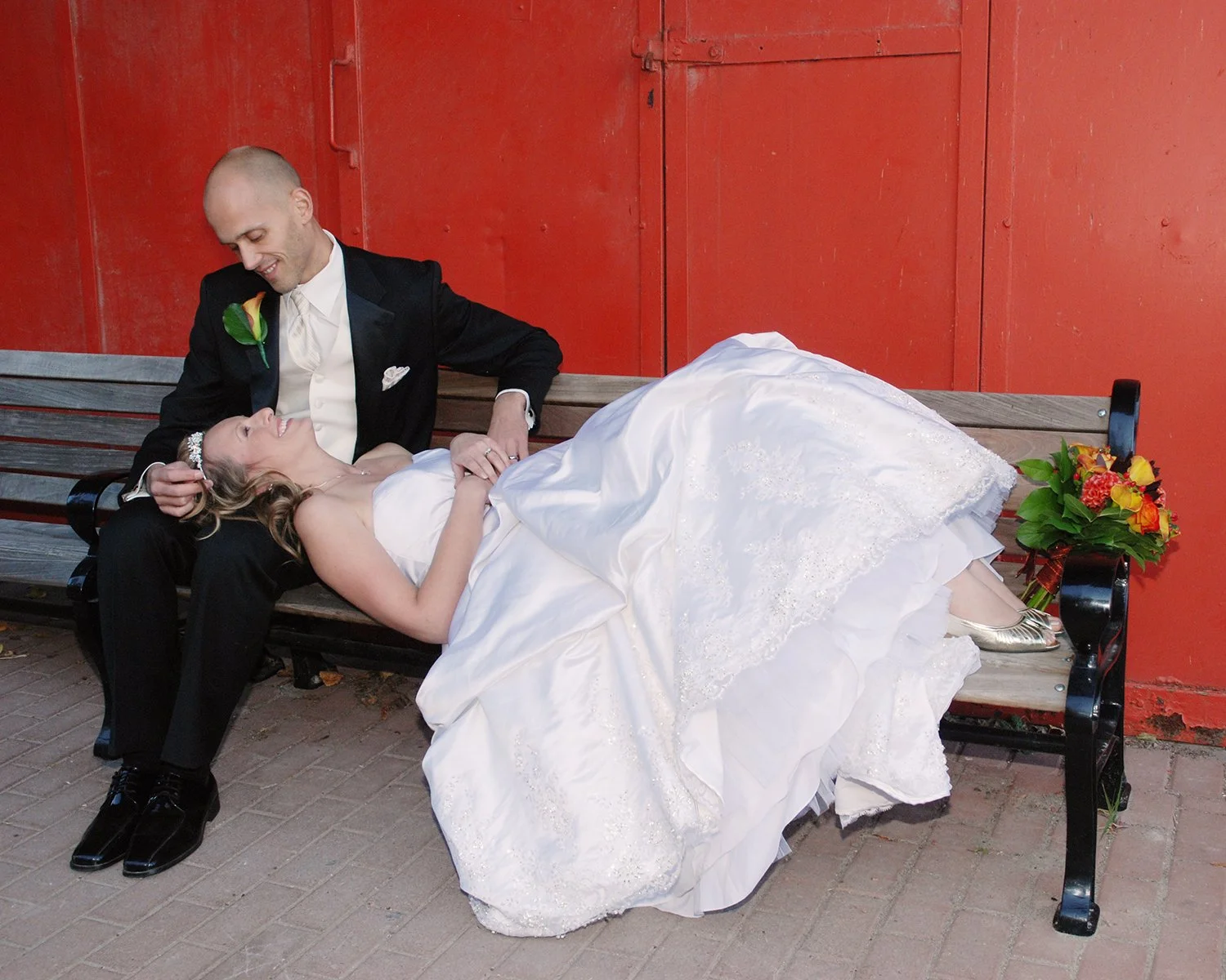 A newlywed couple on a bench with the bride lying down and the groom sitting beside her, smiling and looking at her, with a orange red wall in the background and a bouquet of flowers on the bench.