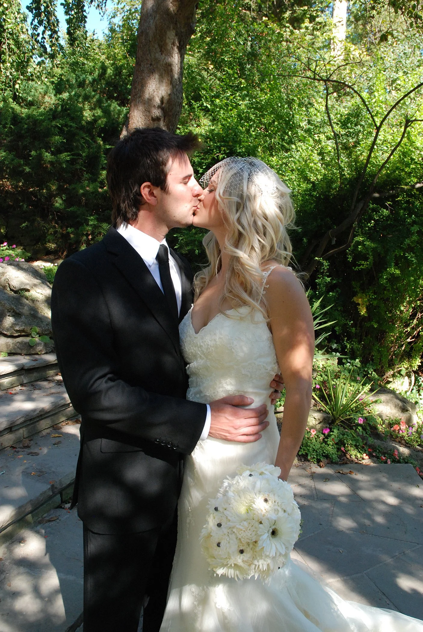 A newlywed couple kissing outdoors surrounded by trees, with the groom in a black suit and the bride in a white wedding dress holding a bouquet of white flowers at Edward Gardens in Toronto Ontario.