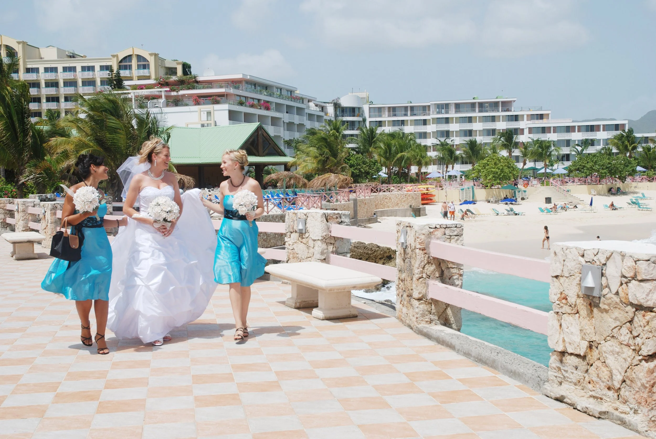 Three women, one in a wedding dress and two in matching blue dresses, holding bouquets, walk on a patio near the beach with hotels, palm trees, in Saint Martin. Destination wedding.