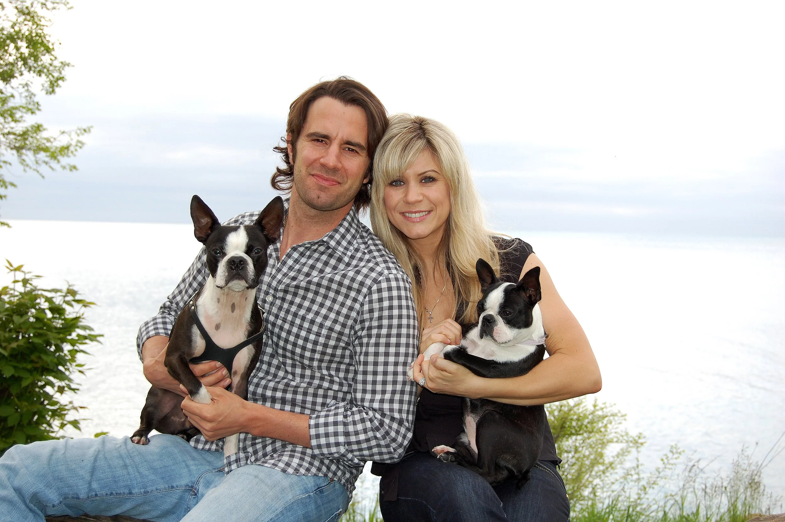 A smiling man and woman sitting outdoors near water, each holding a Boston Terrier dog, with green bushes and a cloudy sky in the background. Engagement photo session in Ajax Ontario.