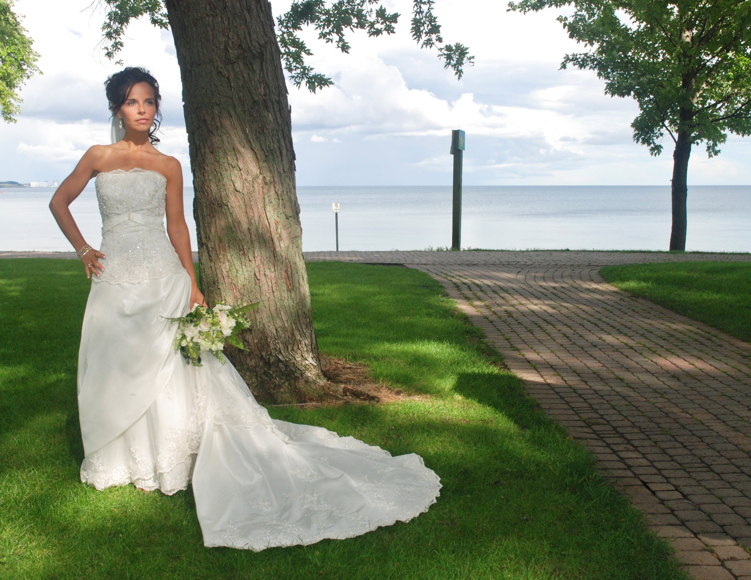 A bride in a white wedding dress holding a bouquet stands next to a tree on a grassy area near a paved walkway, with water and a cloudy sky in the background in August. Oshawa Ontario.