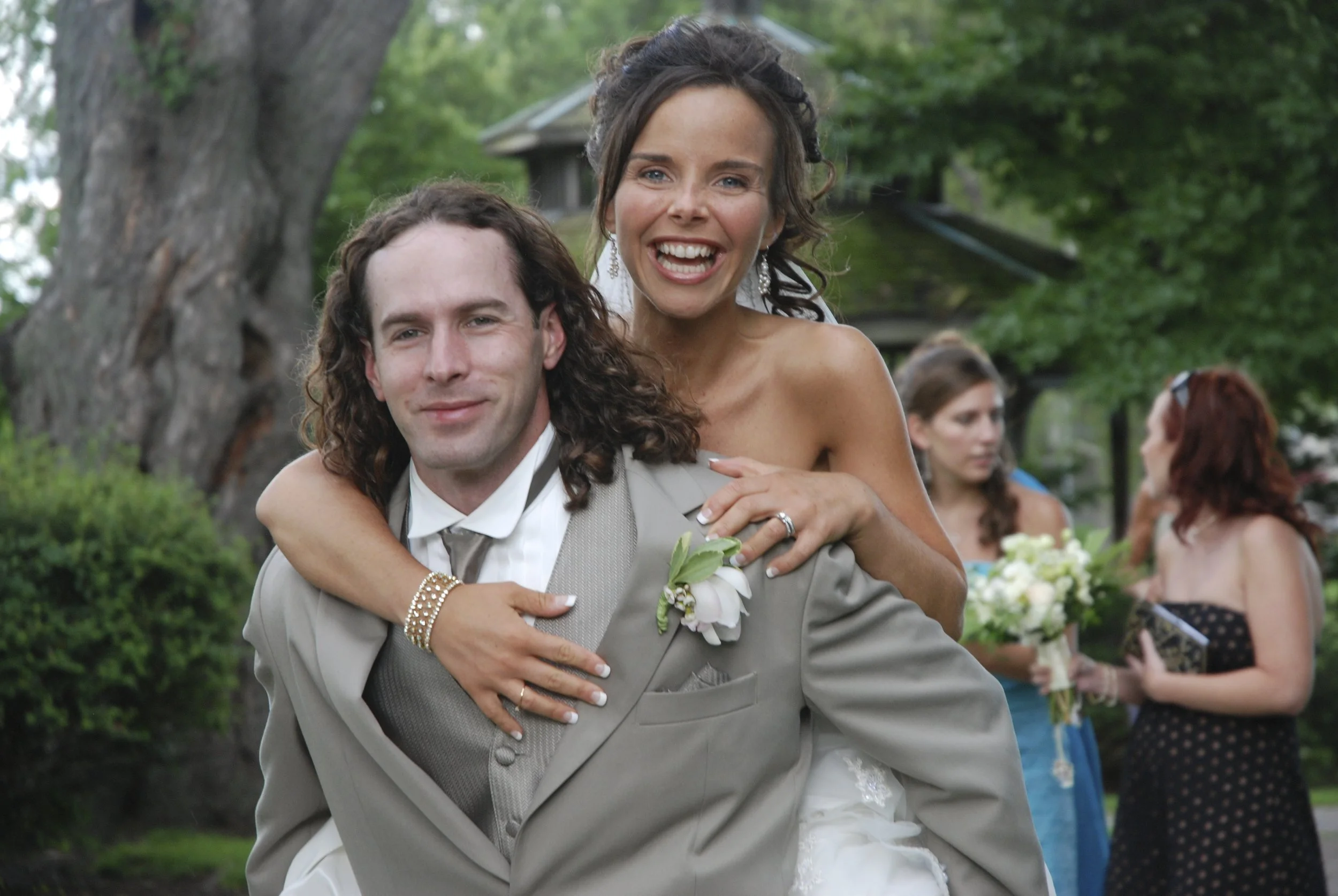 A bride and groom at their wedding, with the bride giving the groom a piggyback ride outdoors among trees and greenery, with other women holding bouquets in the background. Oshawa Ontario.