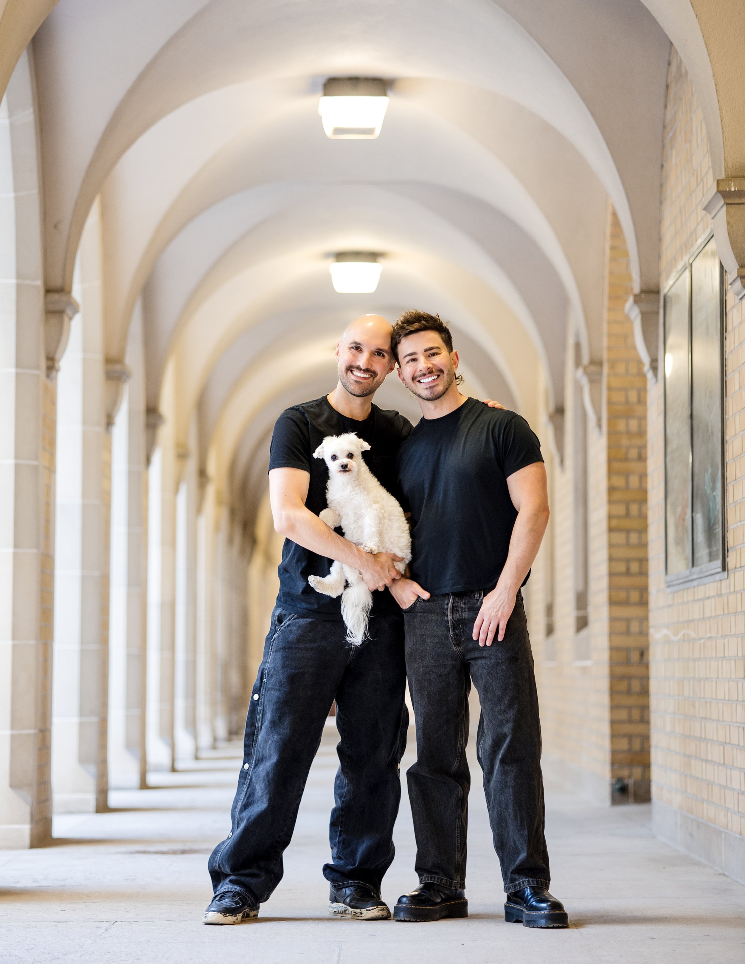 Two gay men standing in an arched outdoor corridor at St.George's University of Toronto campus smiling at the camera. One man is holding a small white dog.