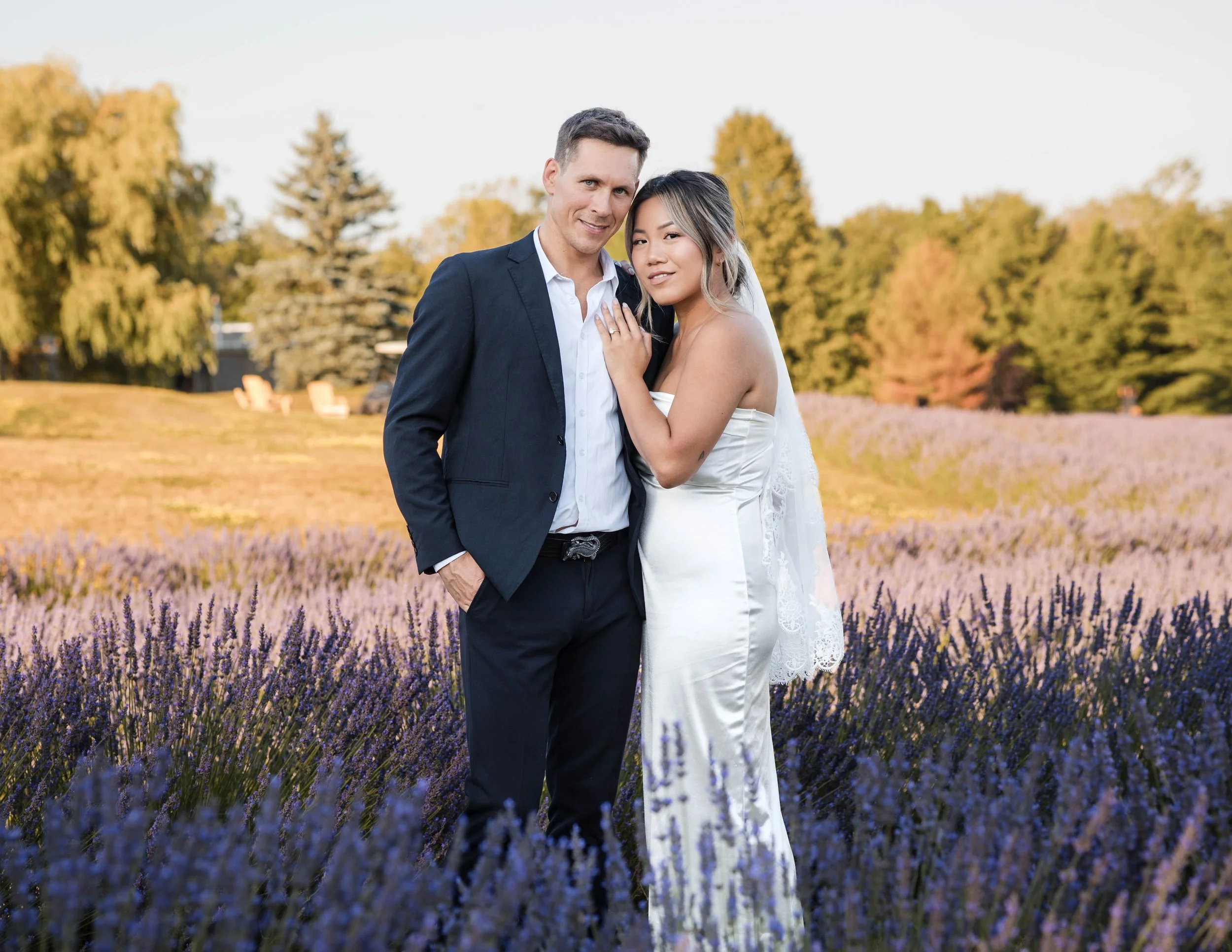 A bride and groom standing close together in a lavender field during sunset, with trees in the background at Tranquility Trails Farm in Pickering Ontario.
