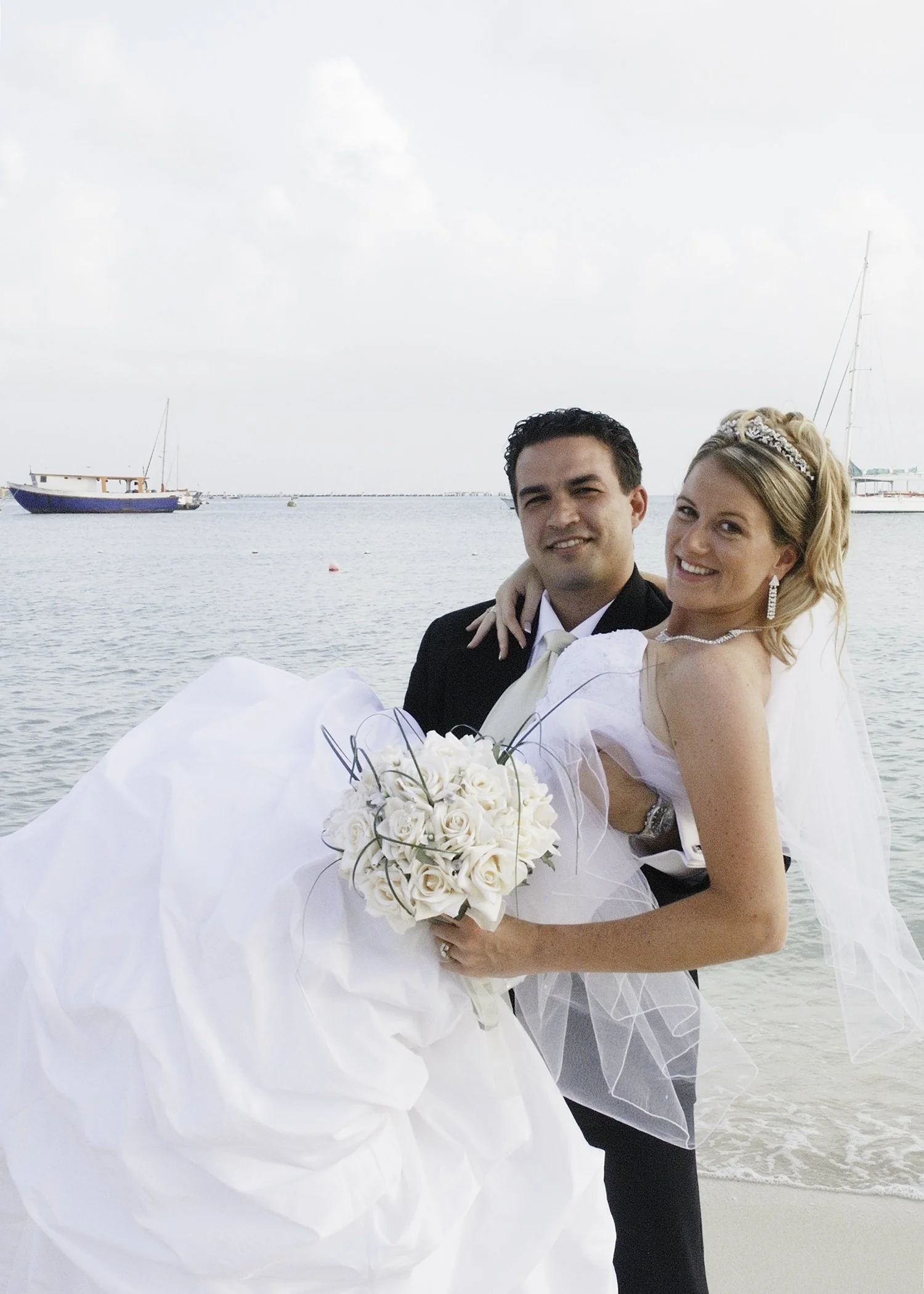 A newly married couple on a beach, with the groom holding the bride and both smiling at the camera. The bride wears a white wedding dress and veil, holding a bouquet of white roses. The background shows calm ocean water and boats, Saint Martin.