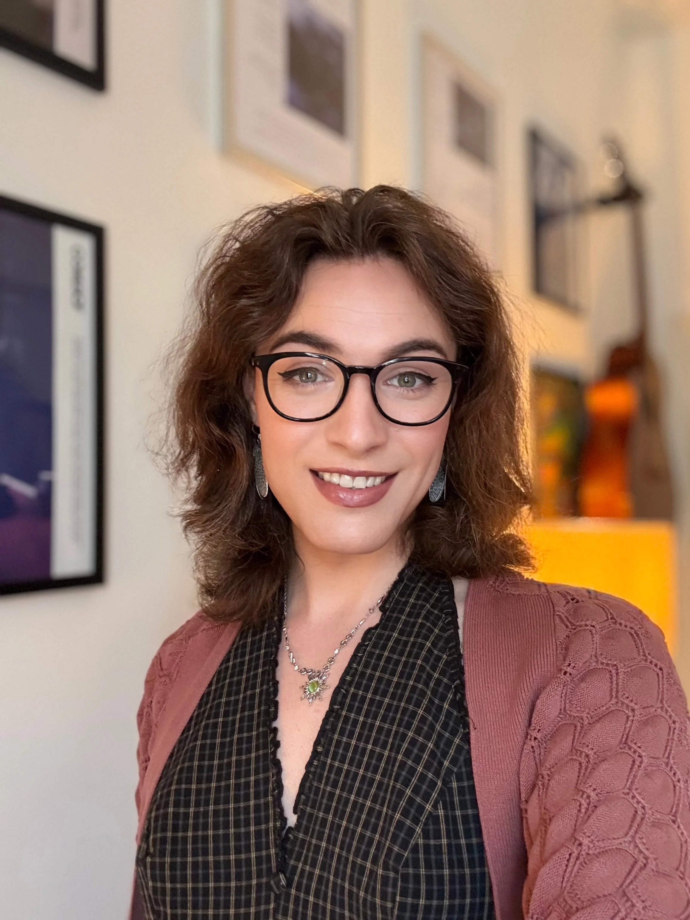 A woman with glasses and curly brown hair smiling at the camera, wearing a pink cardigan over a black and white checkered shirt, with framed pictures on the wall behind her.