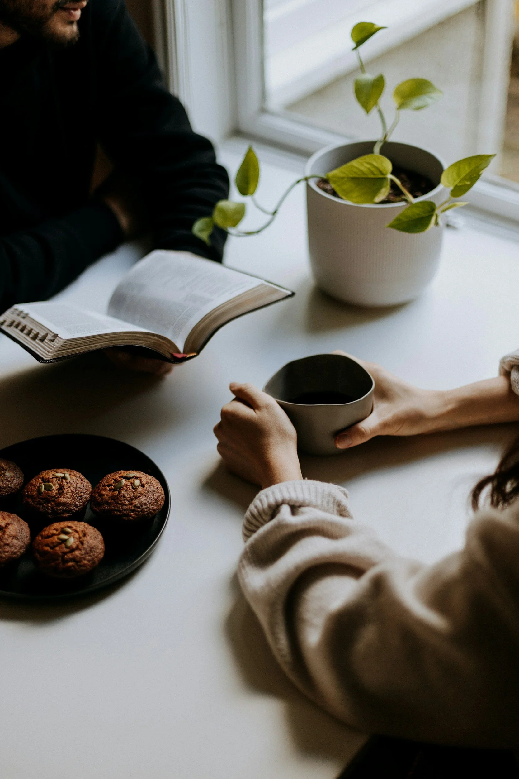 Two people sitting at a white table, one holding a cup of coffee or tea, the other with an open book, with a plate of muffins and a potted plant on the table.