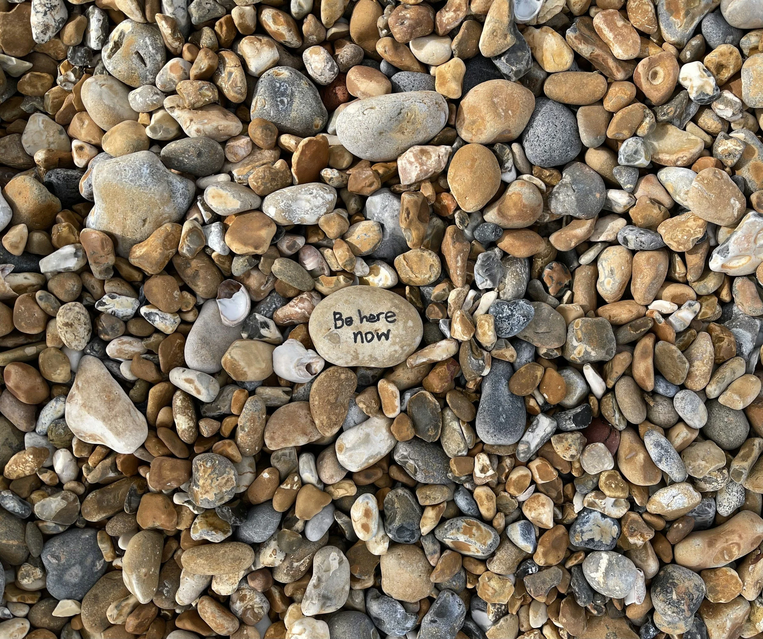 A bed of small, smooth, multicolored pebbles with a single beige pebble that has the words 'Be here now' written on it.
