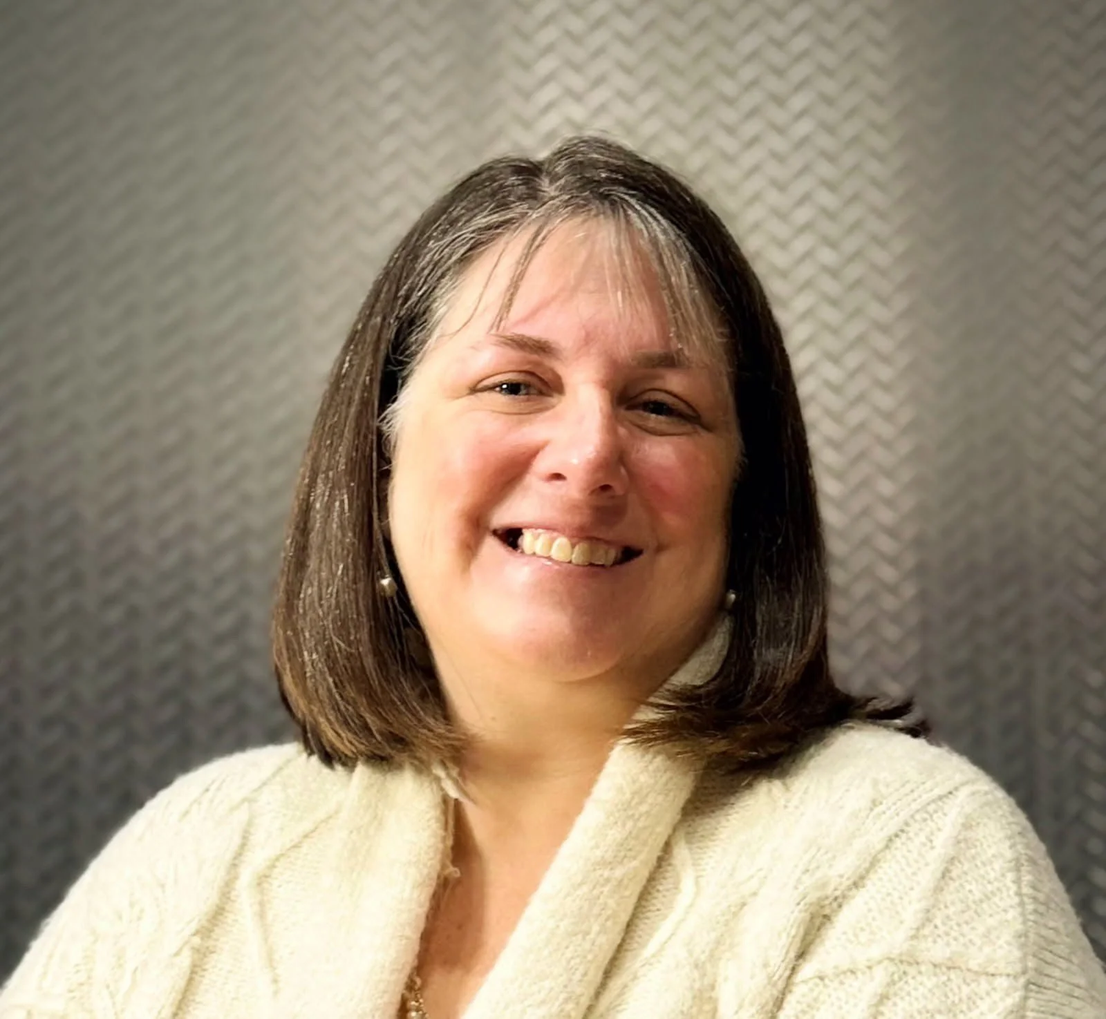A woman with shoulder-length brown hair, smiling, wearing a cream-colored sweater and pearl earrings, standing in front of a textured wallpaper background.