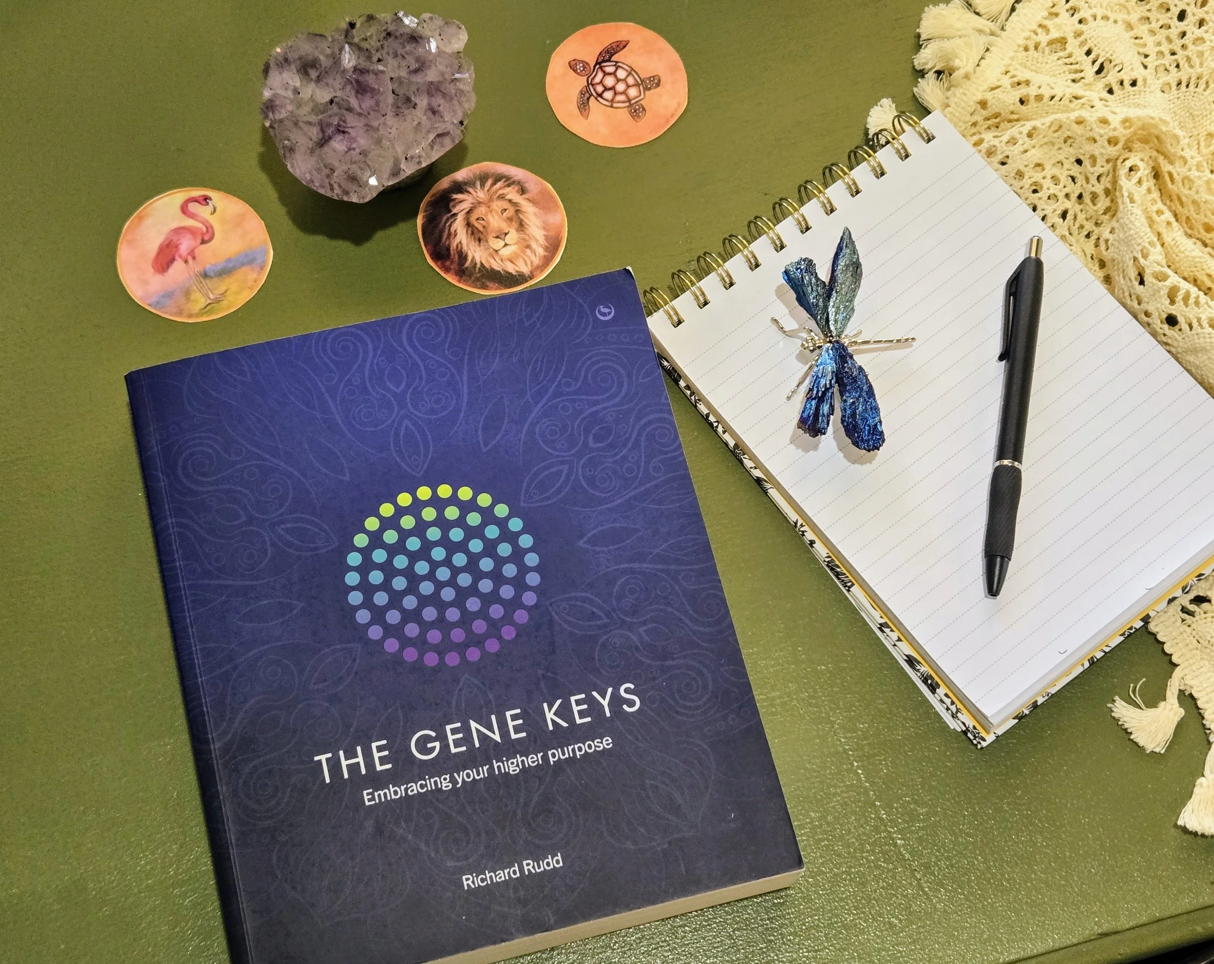 A book titled 'The Gene Keys: Embracing your higher purpose' by Richard Rudd, a spiral notebook with a pen, a decorative butterfly on the notebook, various animal-themed stickers including a flamingo, lion, turtle, and a heart-shaped crystal, a yellow knitted cloth, all on a green surface.