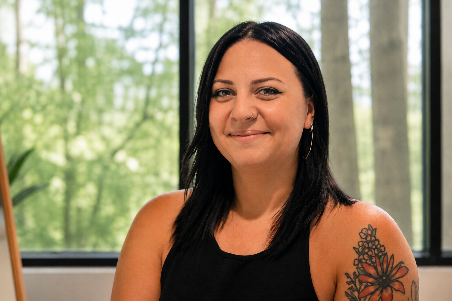 A woman with black hair, wearing a black sleeveless top, gold hoop earrings, and a floral tattoo on her upper arm, smiling at the camera inside a room with large windows and green trees outside.