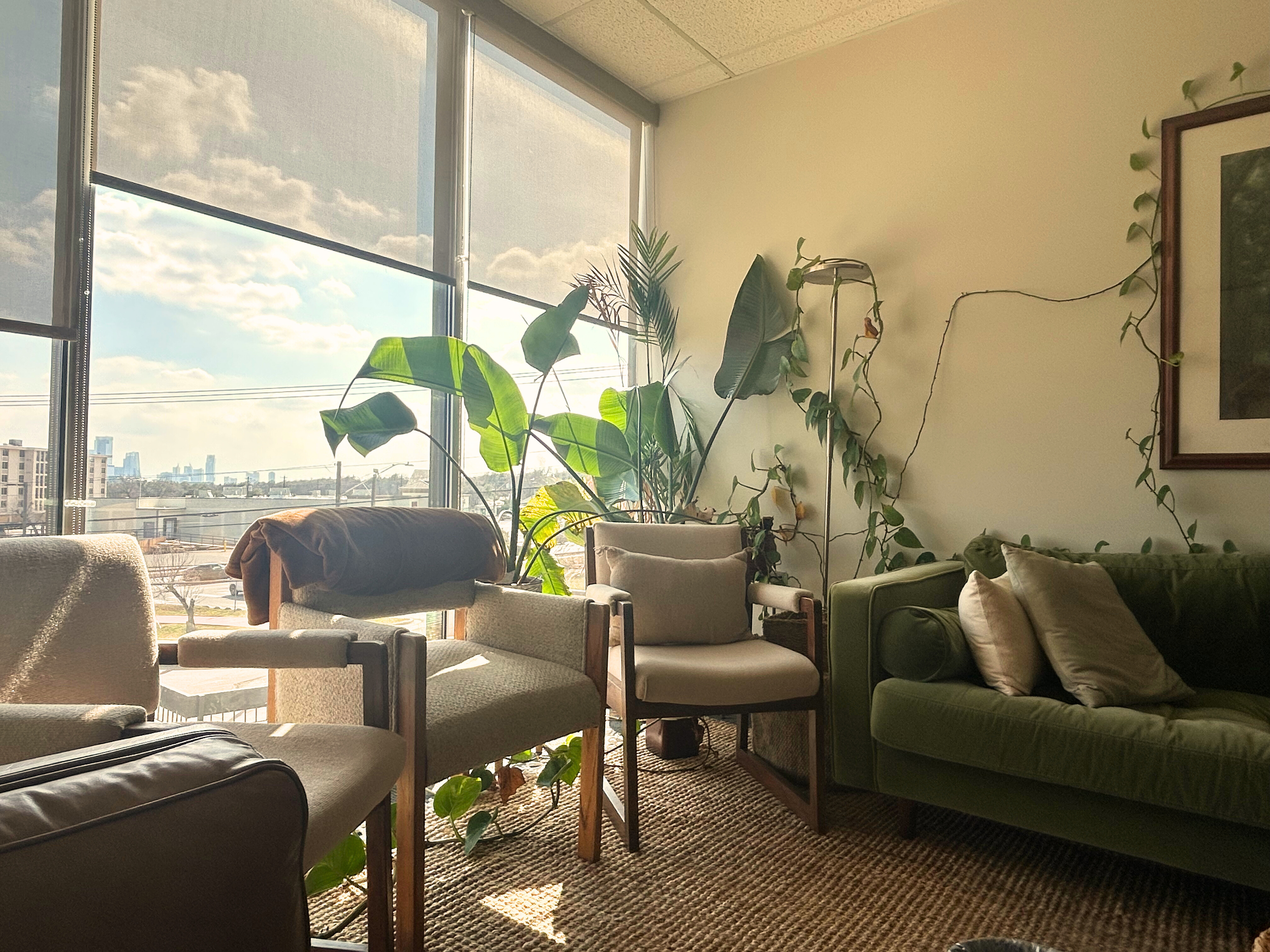 Dietitian office space for in person nutrition sessions with large windows, various green plants, a beige armchair, a green sofa with white pillows, and a framed picture on the wall.