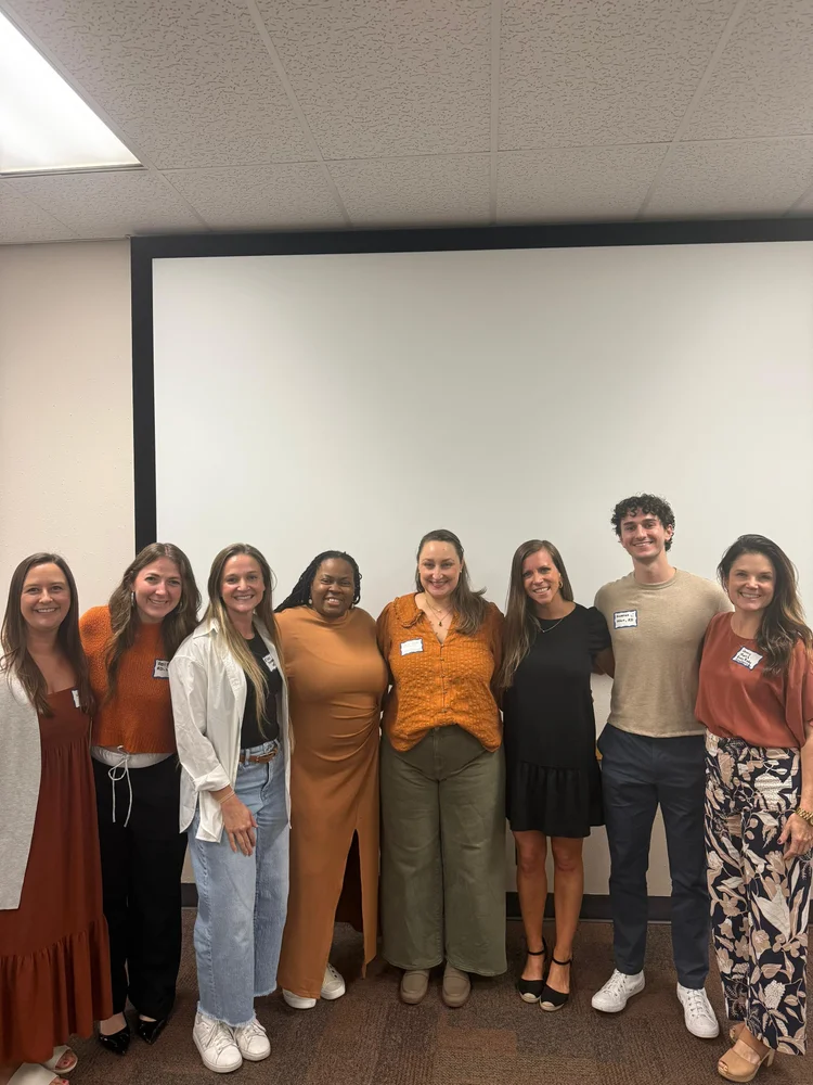 Eight people standing in a row in front of a projection screen, most wearing rust and orange tones, with name tags on their shirts.