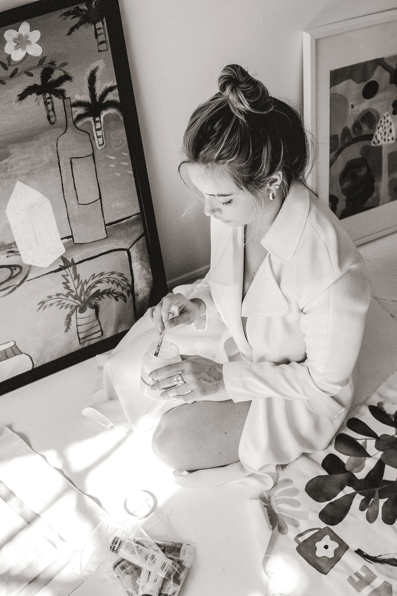 A woman sitting on a bed in a white outfit, mixing something in a small jar, with colorful artwork and a framed picture of a beach scene in the background.