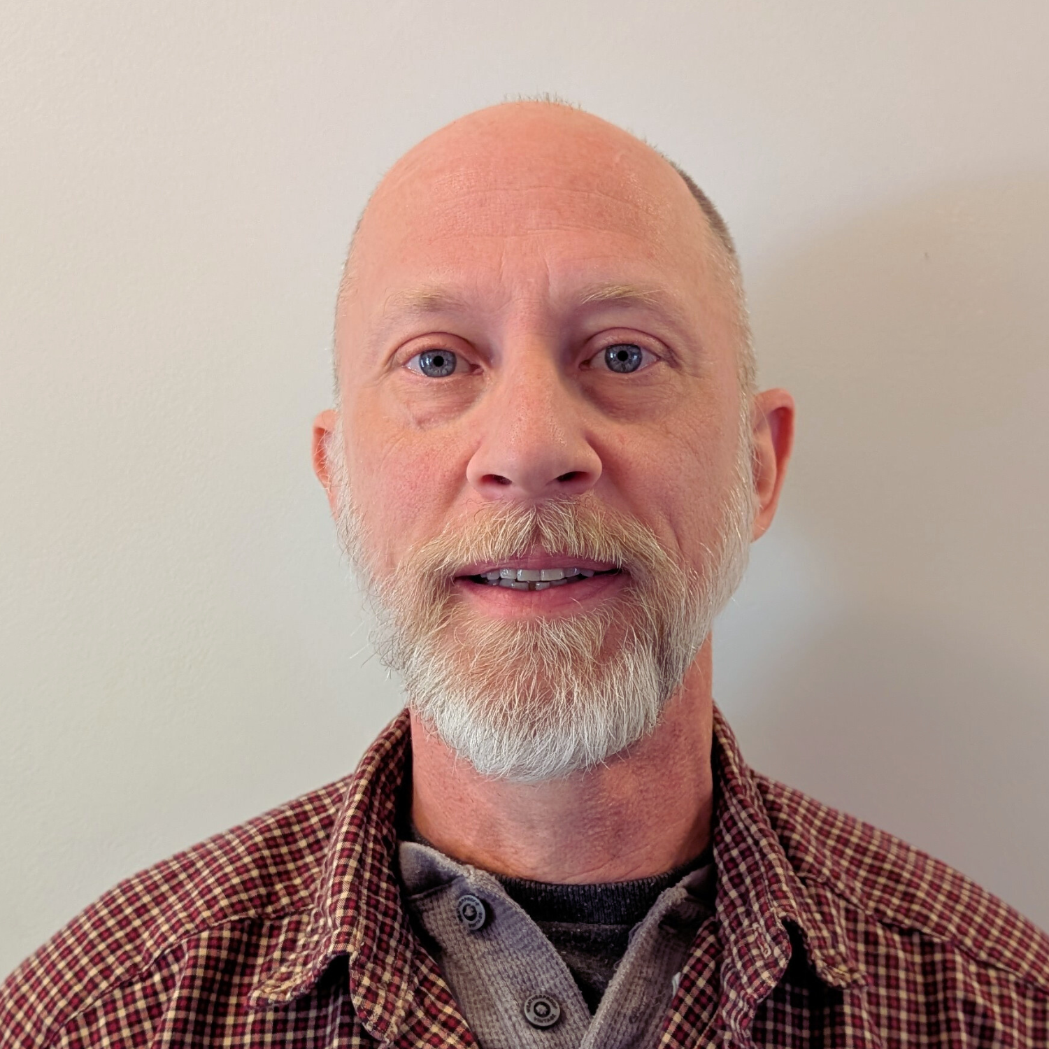 A middle-aged man with blue eyes, a beard, and a mustache, wearing a plaid shirt over a gray undershirt, standing against a plain white wall.