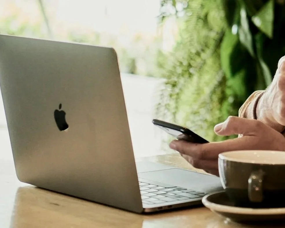 Person using a smartphone next to a silver MacBook laptop with a black Apple logo on a wooden table, with a coffee cup nearby, in a bright setting with greenery outside the window.