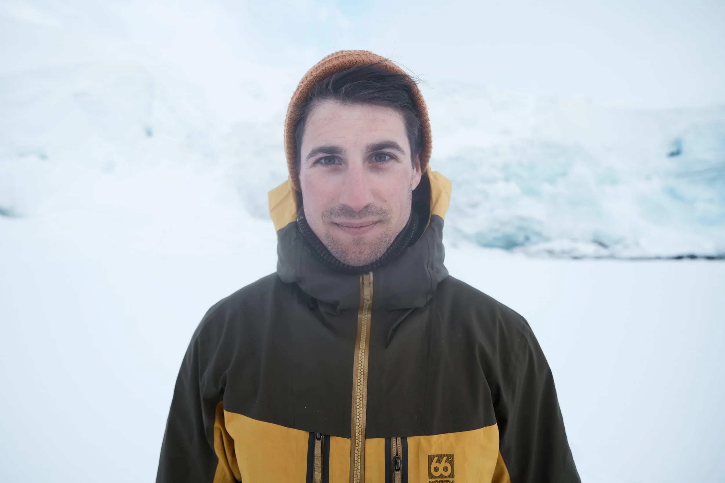 A young man with dark hair and blue eyes wearing a brown beanie and a black and yellow winter jacket, standing outdoors in a snowy environment.