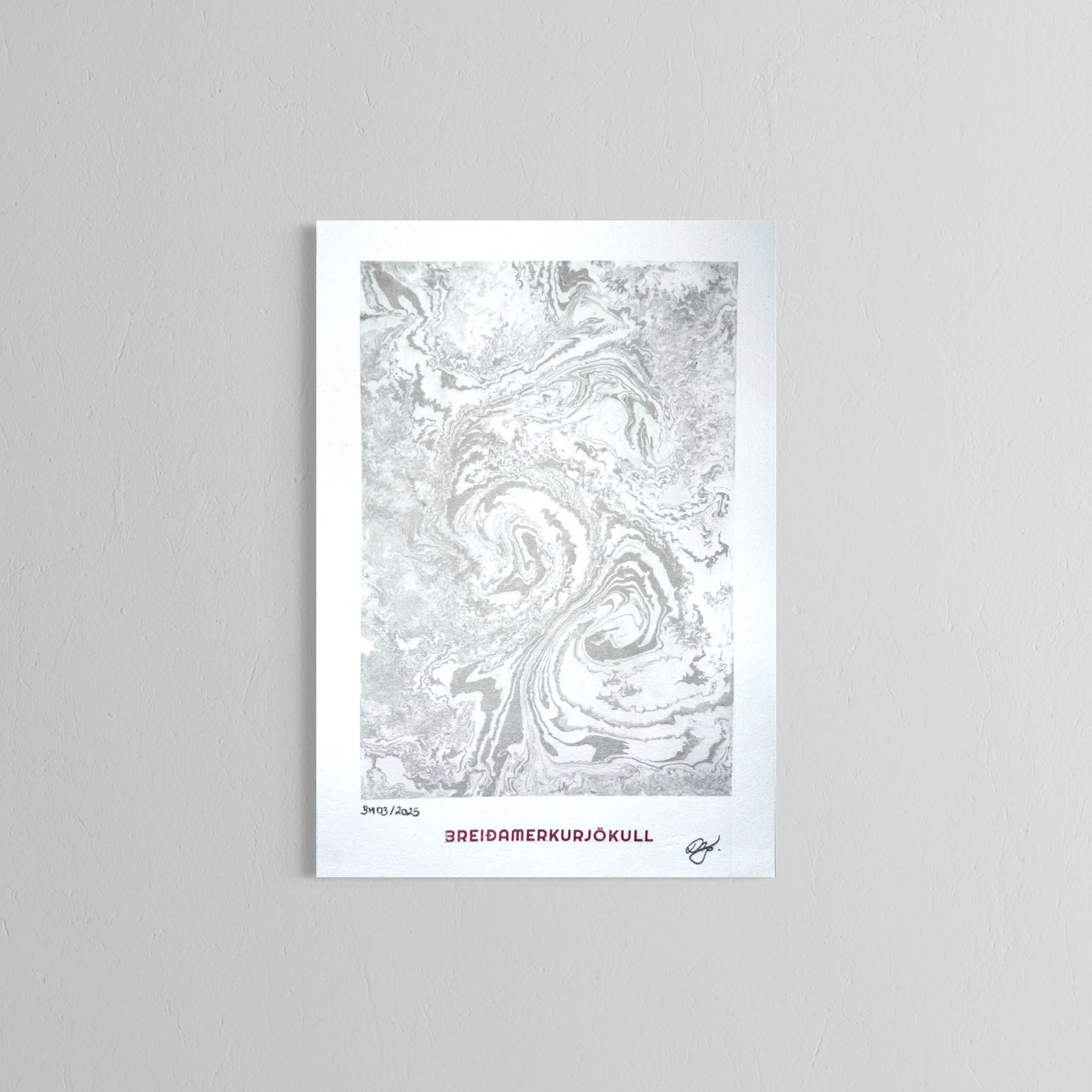 A handmade print with a marbled black and white image, handwritten date, title in red reading 'Breidamerkurjokull,' and a signature, placed on a plain white wall.