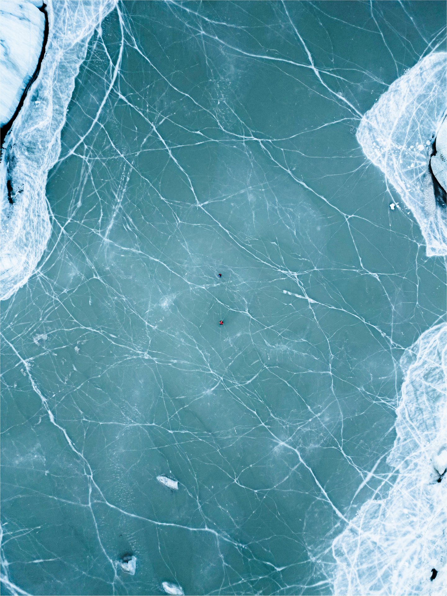 Aerial view of two kayakers on a frozen icy lake with cracks and snow-covered edges.