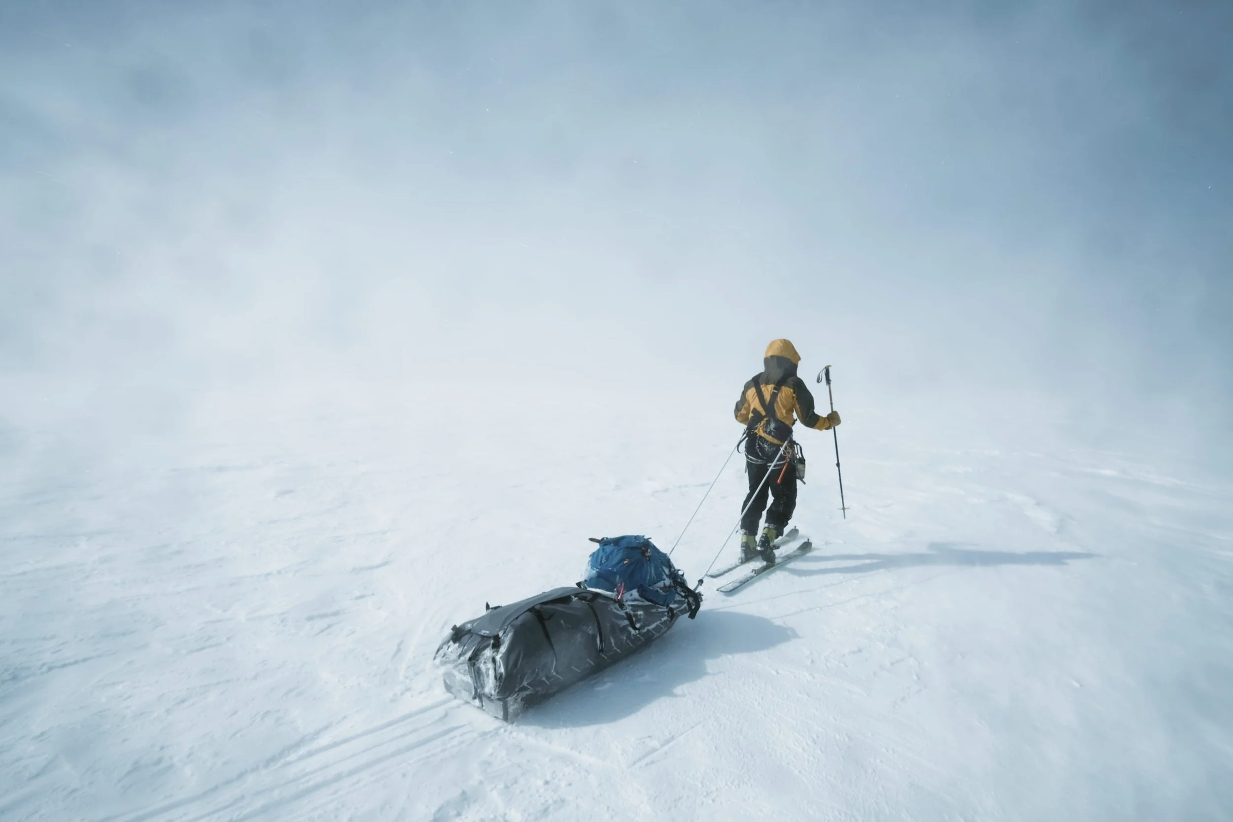 A person in yellow winter gear and a helmet is skiing on snow, pulling a sled with gear, in a foggy, snowy landscape.