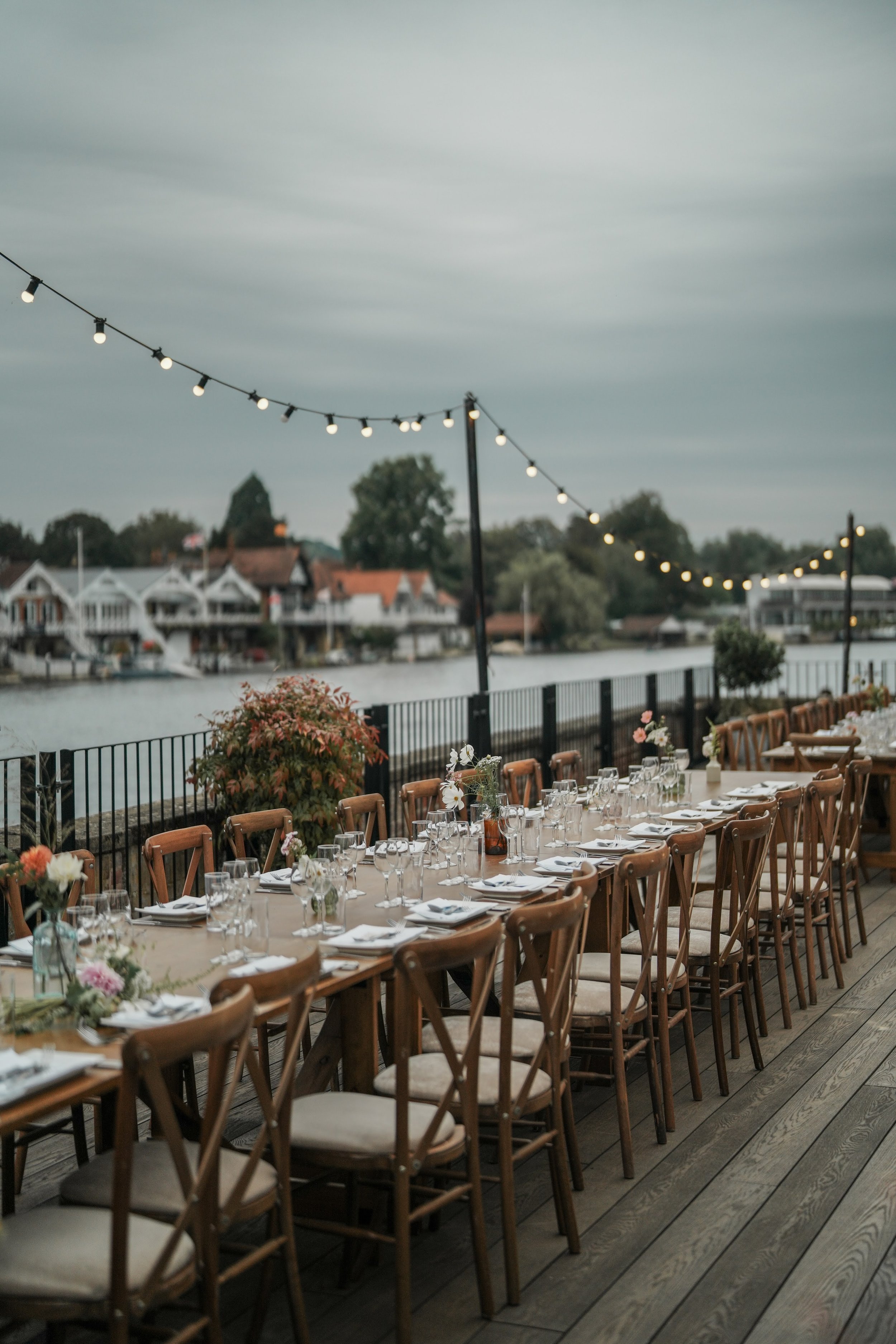 Outdoor dining setup with long wooden tables, chairs, and floral centerpieces, overlooking a river under string lights and cloudy sky.