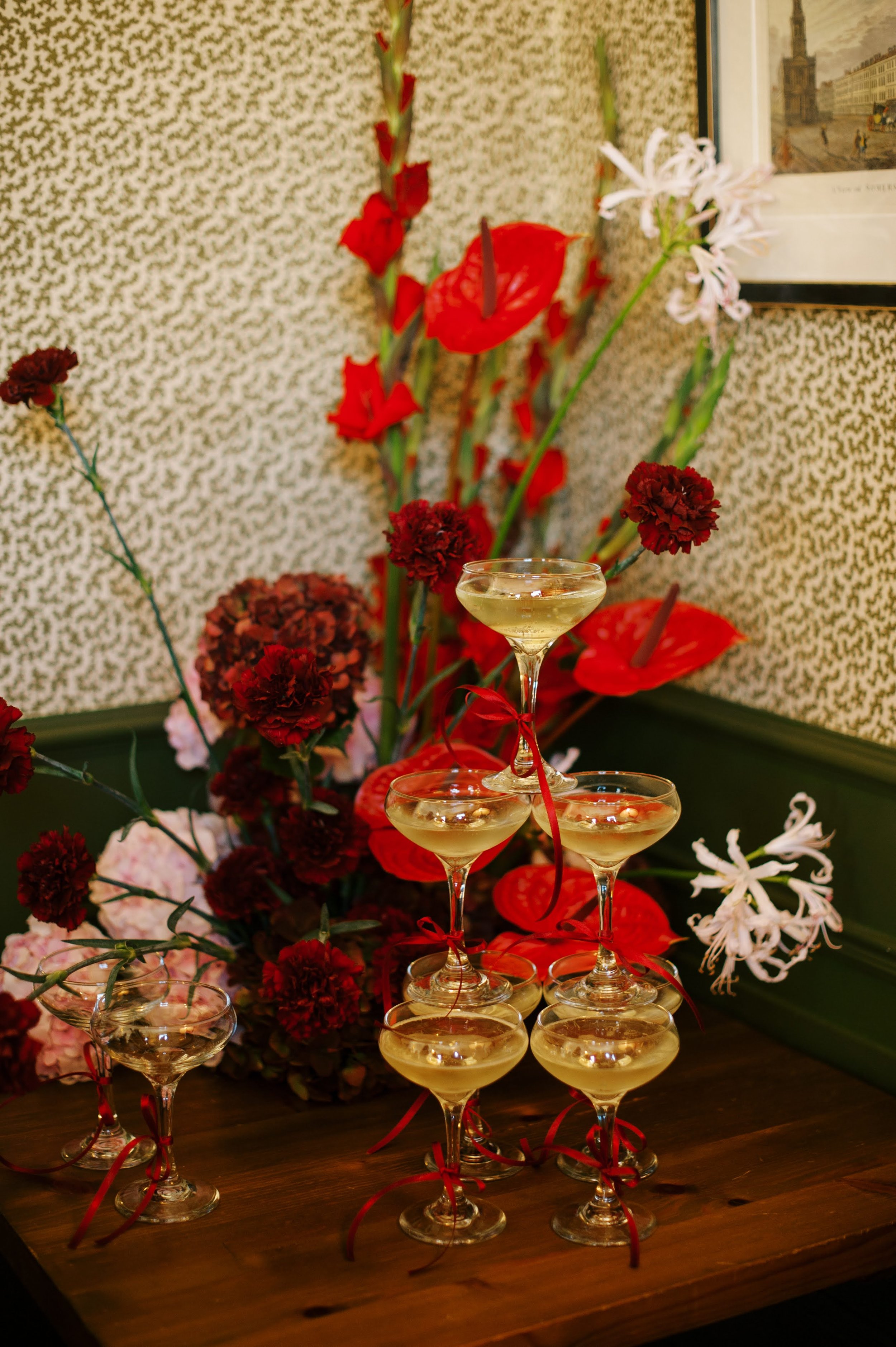 A pyramid of six cocktail glasses filled with a light-colored beverage, decorated with red ribbons, placed on a wooden surface. Behind the glasses, there is a floral arrangement with large red, pink, and white flowers, set against a patterned wall.