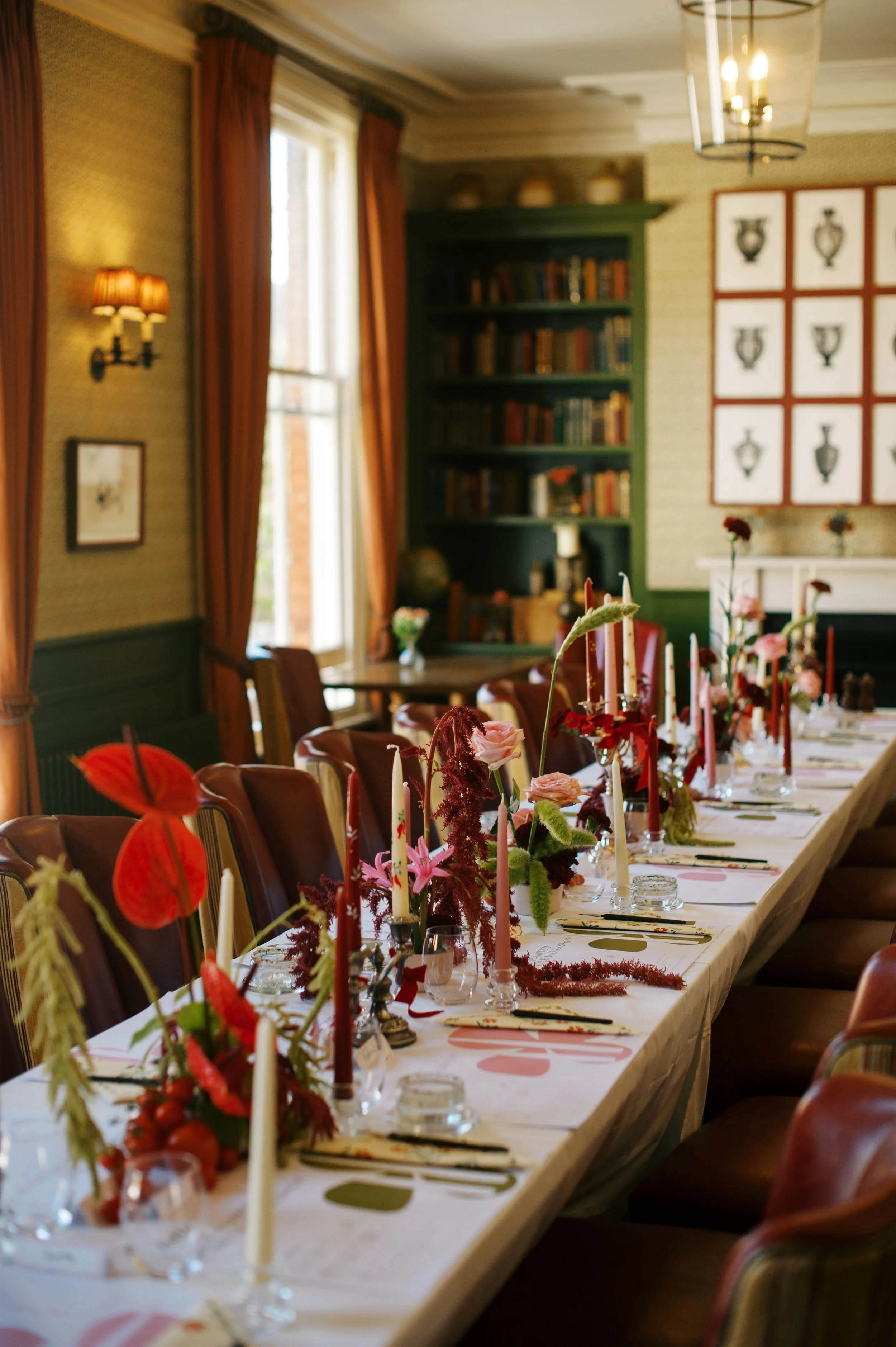 Decorated dining table with flowers and candles in a warmly lit room with bookshelves and framed artwork.