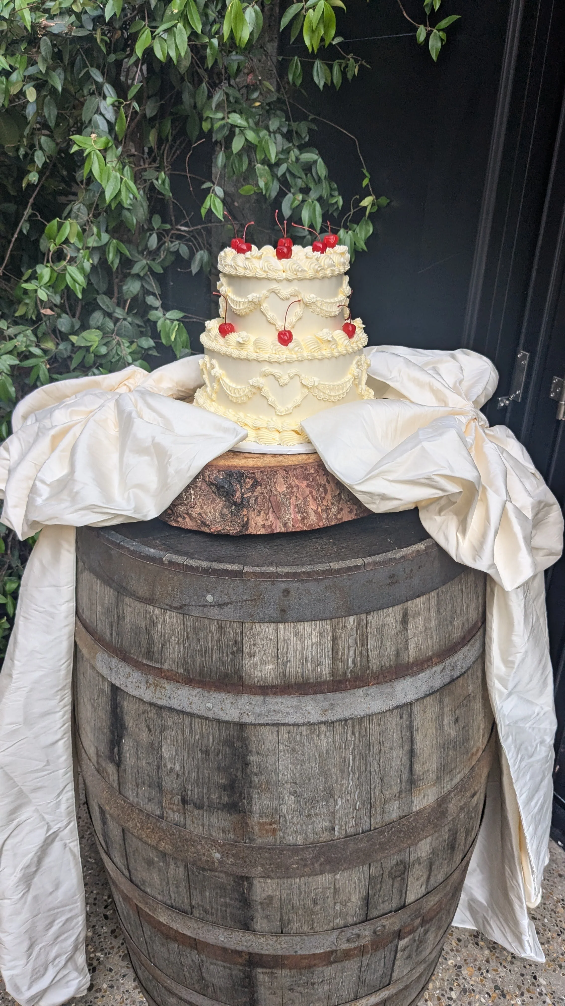 A three-layer white wedding cake decorated with cream swirls and cherries on top, placed on a wooden barrel with white fabric draped around it, outdoors with green foliage in the background.