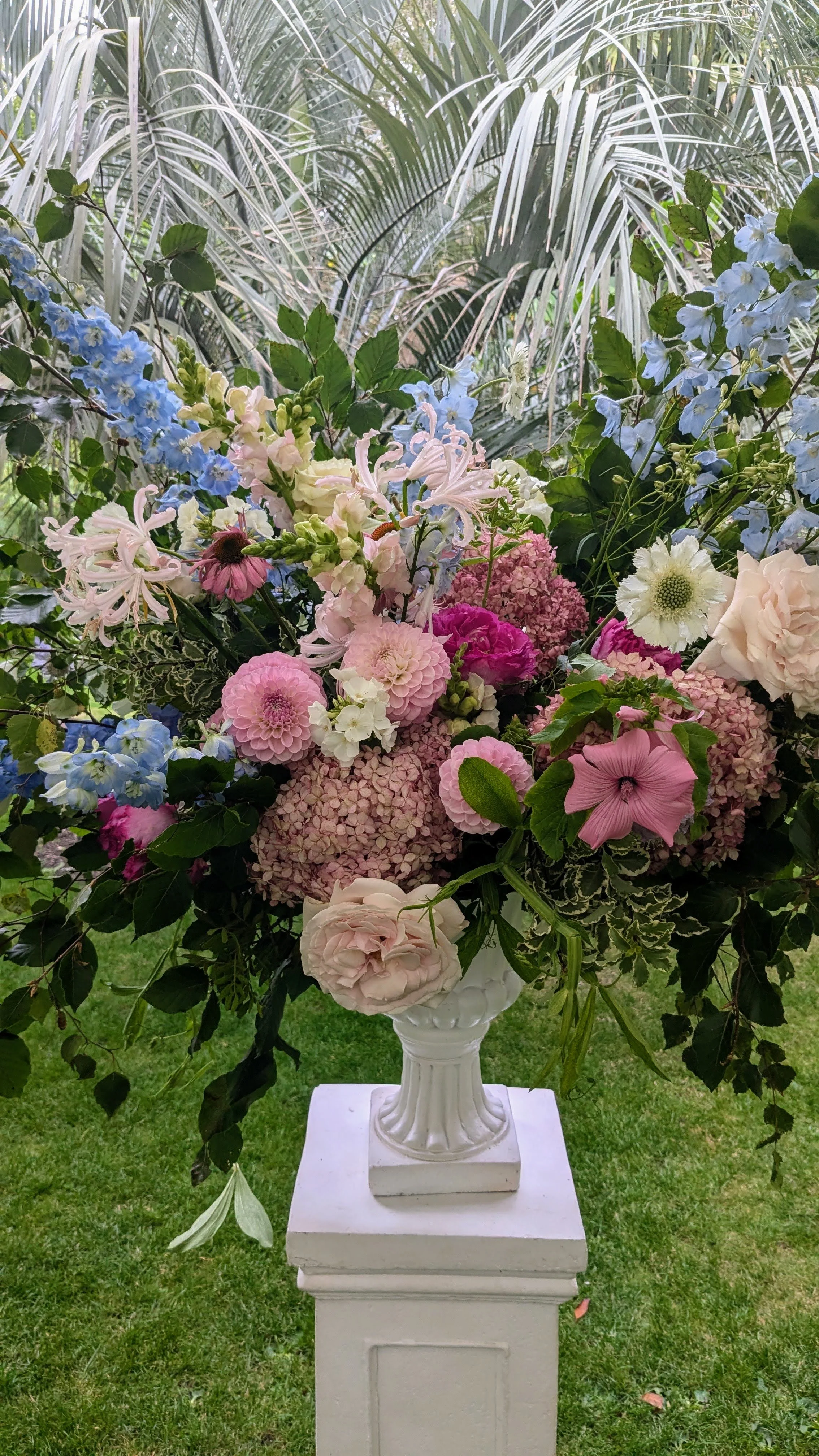 A large bouquet of pink, white, and blue flowers in a white vase on a pedestal, with green grass and lush foliage in the background.