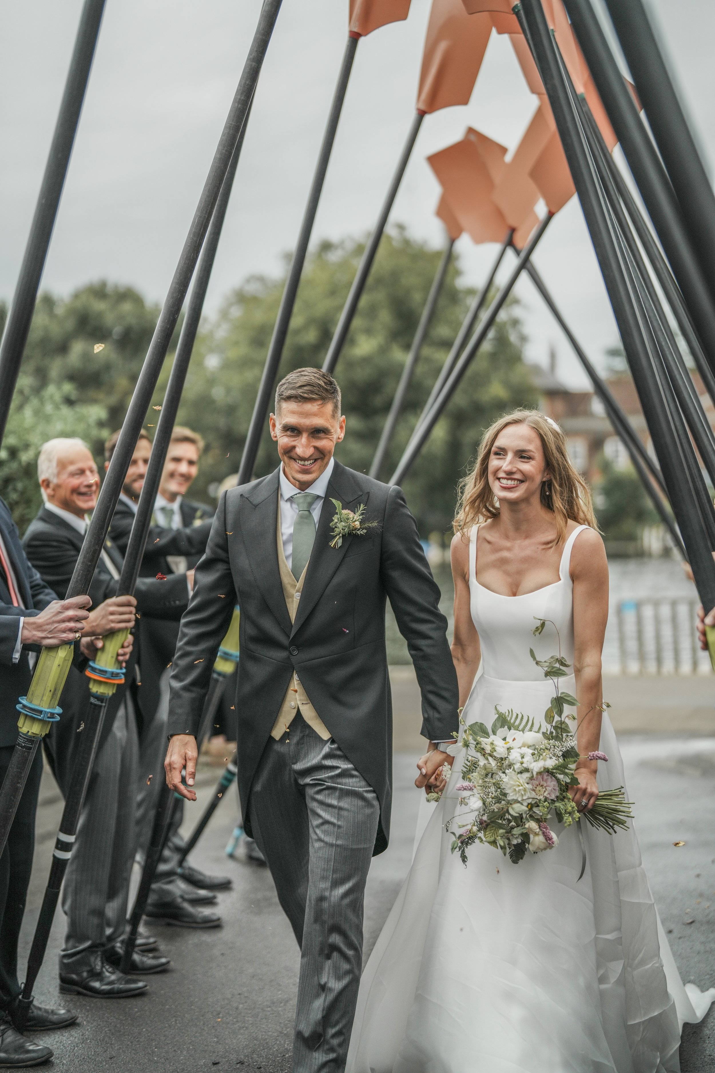 A bride and groom smiling as they walk hand in hand through a celebration, surrounded by wedding guests holding large dip poles, outdoors near a waterway.