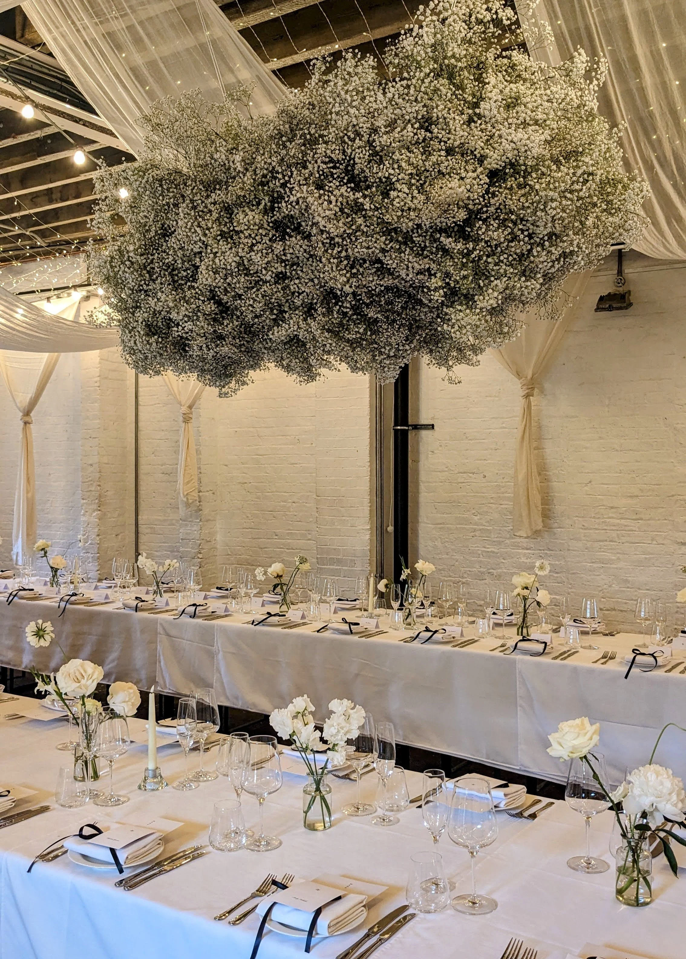 Elegant banquet hall decorated with white flowers, hanging floral installation, and long tables with white tablecloths, glassware, and napkins tied with black ribbons for a formal event.
