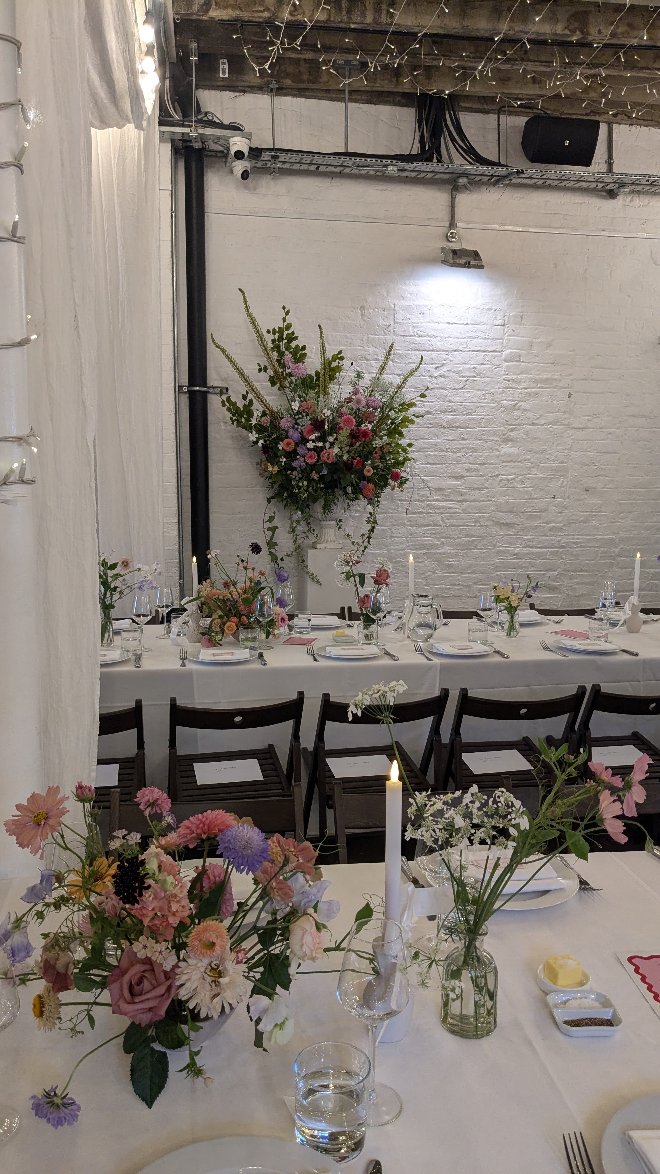 A decorated banquet table with floral centerpieces, candles, and place settings, set in a rustic venue with white brick walls and string lights.
