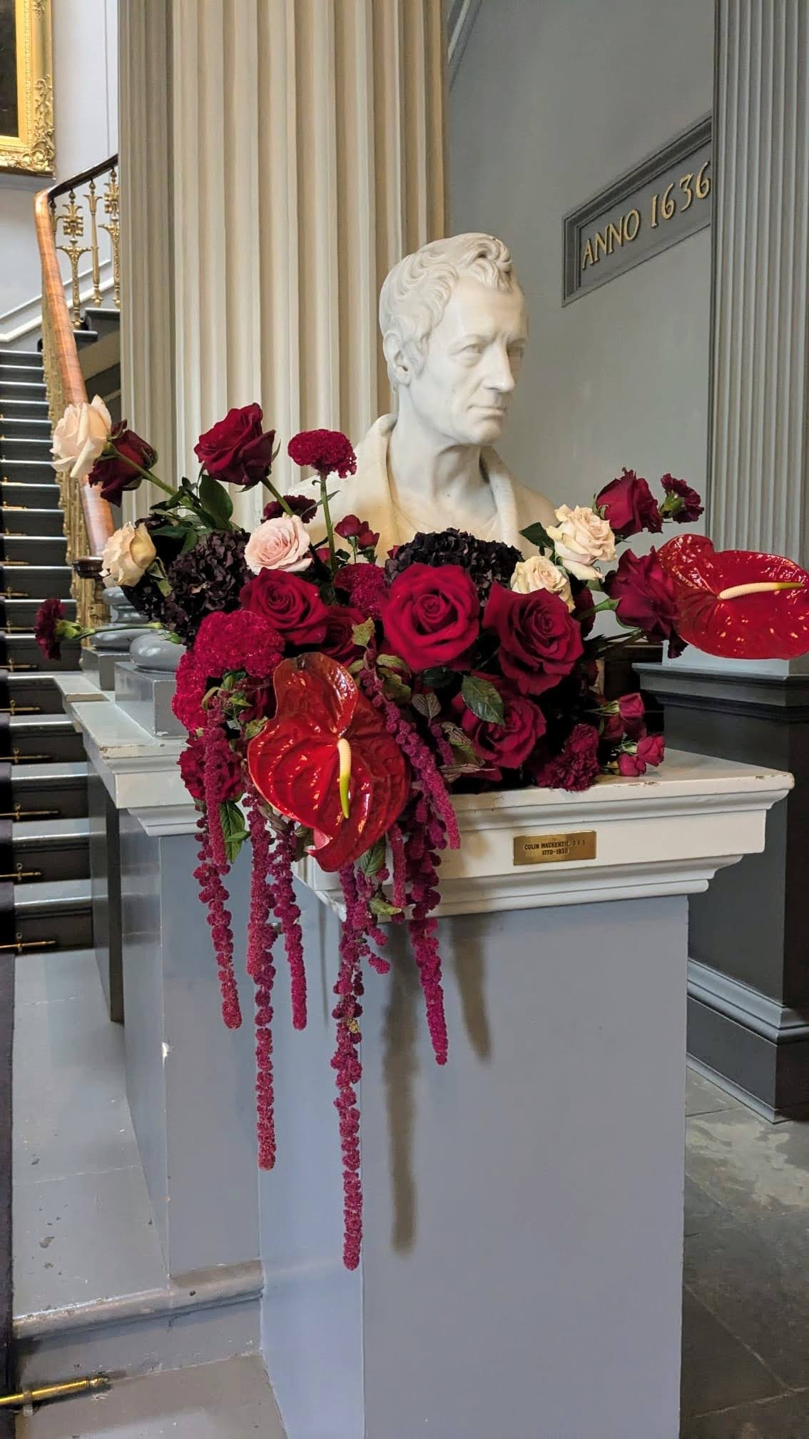 Statue of a man’s bust decorated with red, pink, and dark flowers and red anthuriums, placed on a pedestal inside a building with gray walls and a staircase.