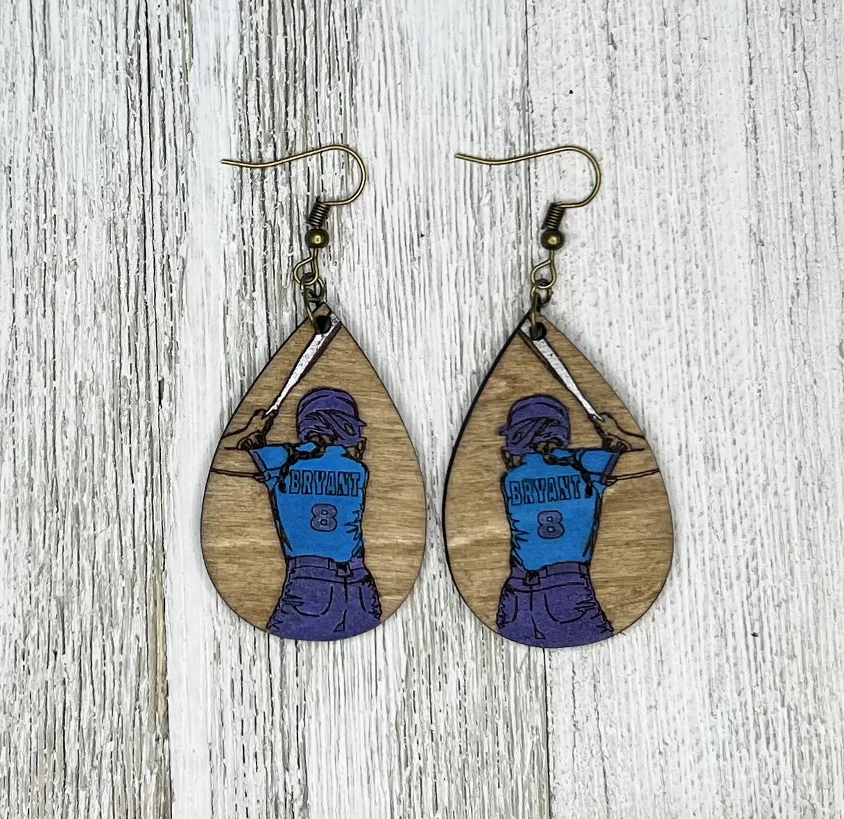 Wooden earrings with a drawing of a person from behind, wearing a blue jersey with the word "BRYANT" and the number 8, holding a tennis racket.