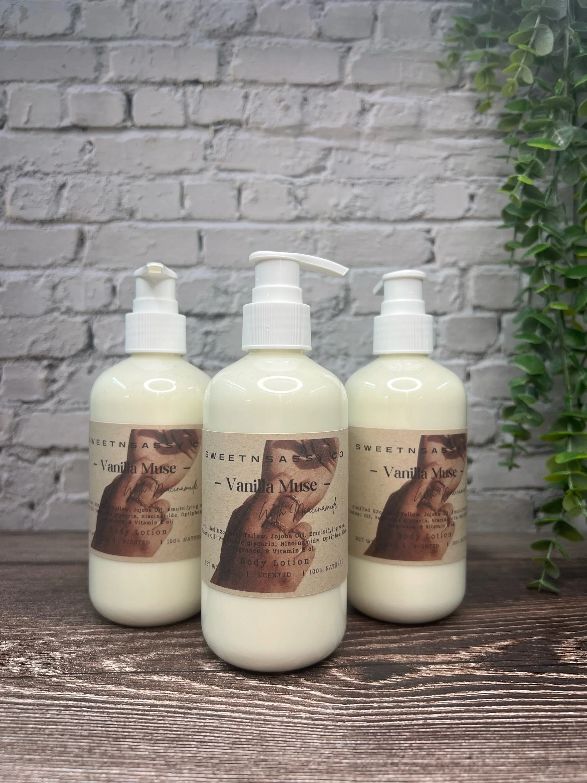 Three bottles of Vanilla Muse body lotion with pump dispensers placed on a wooden surface against a white brick wall with a green plant on the right side.