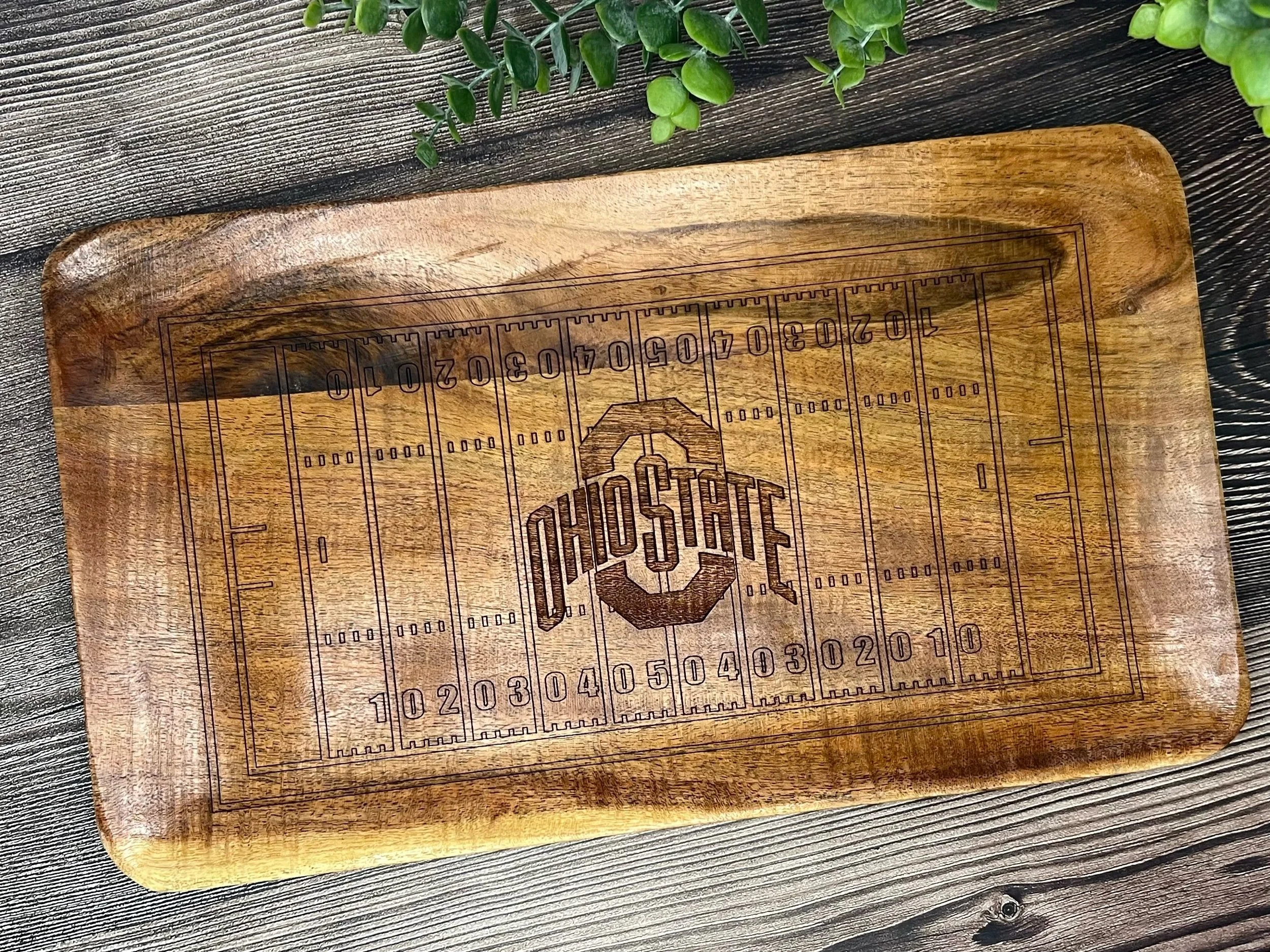 Wooden cutting board with a pizza diagram and a black marker ruler, placed on a dark wood surface with green plants in the background.