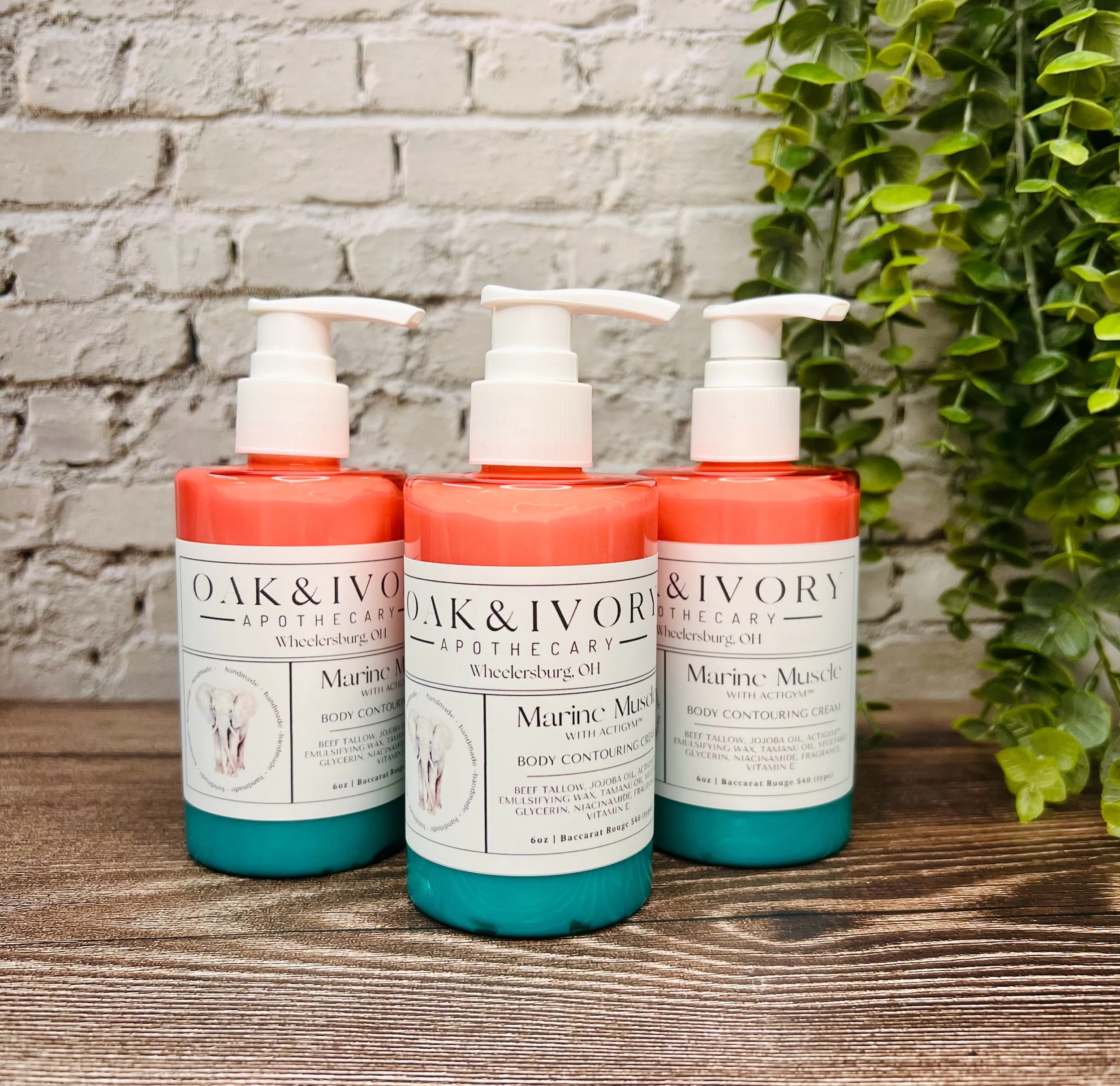 Three bottles of OAK & IVORY body contouring cream in teal, coral, and orange, placed on a wooden surface with a brick wall and green plant in the background.