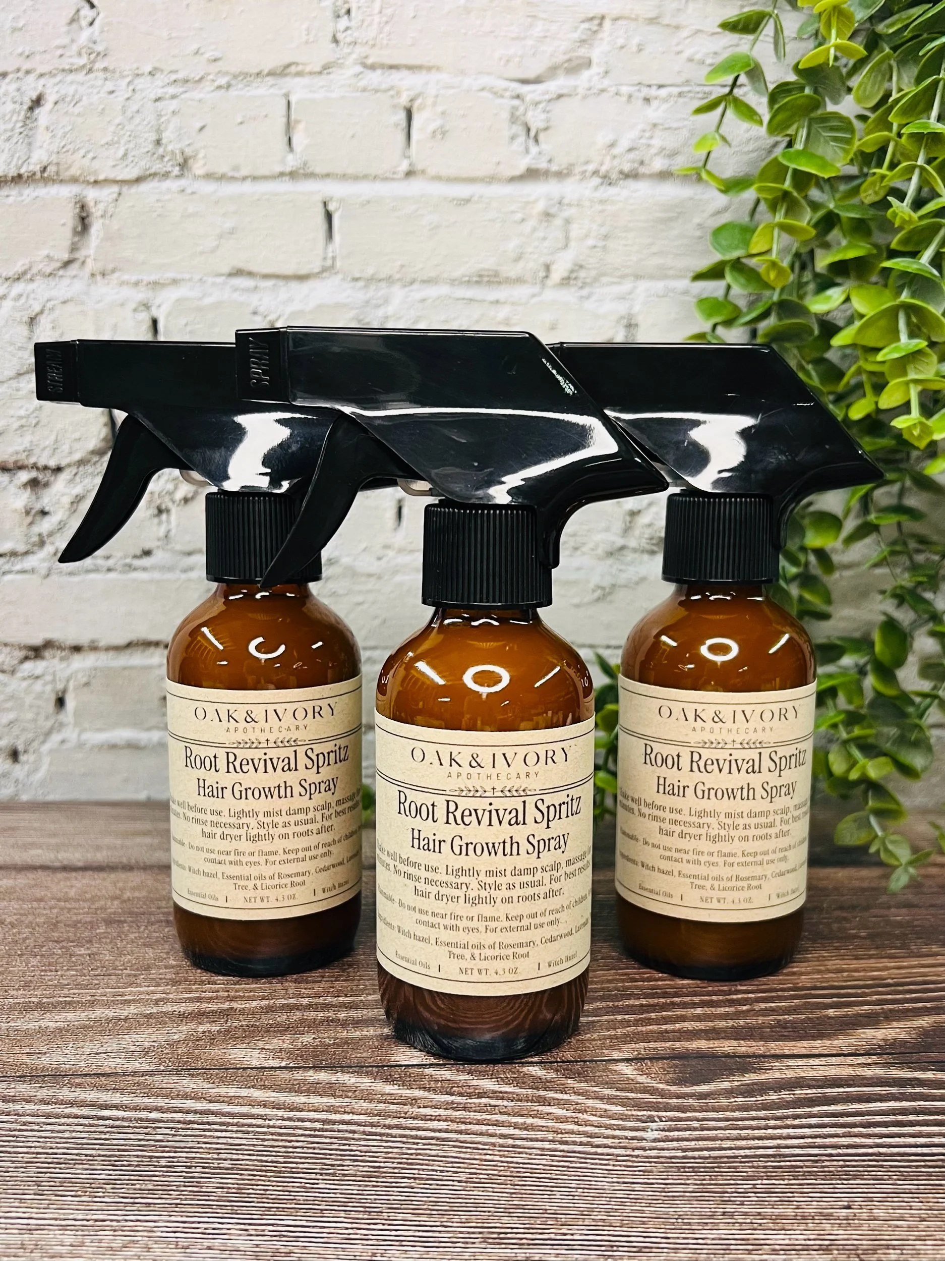 Three amber bottles labeled 'Oak & Ivory Apothecary Root Revival Spritz Hair Growth Spray' with black spray nozzles, placed on a wooden surface with a white painted brick wall and green foliage in the background.