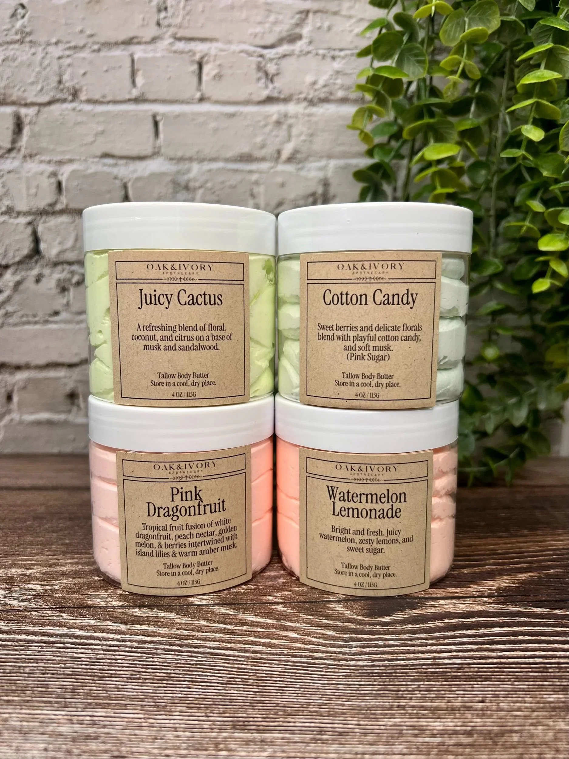 Oak & Ivory Apothecary stacked jars of body butter with labels for various scents including Juicy Cactus, Cotton Candy, Pink Dragonfruit, and Watermelon Lemonade on a wooden surface, with a white brick wall and green plant in the background.