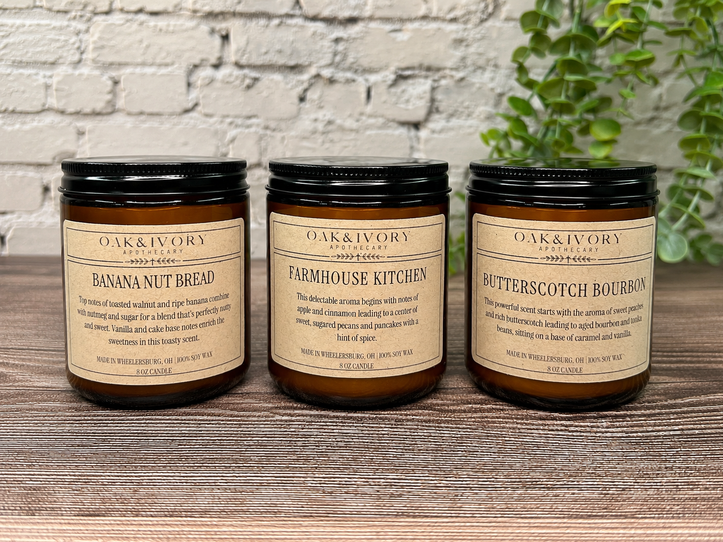Three Oak & Ivory Apothecary soy candles in dark amber jars with black lids, labeled "Banana Nut Bread," "Farmhouse Kitchen," and "Butterscotch Bourbon," are placed on a rustic wooden surface with a white brick wall and green plant in the background.