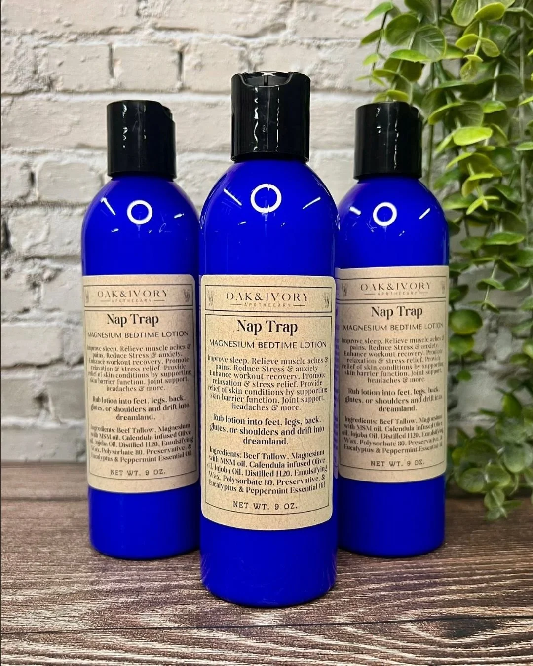 Three blue bottles of Oak & Ivory Apothecary Nap Trap Magnesium Bedtime Lotion on a wooden surface with a white brick wall and green plant in the background.