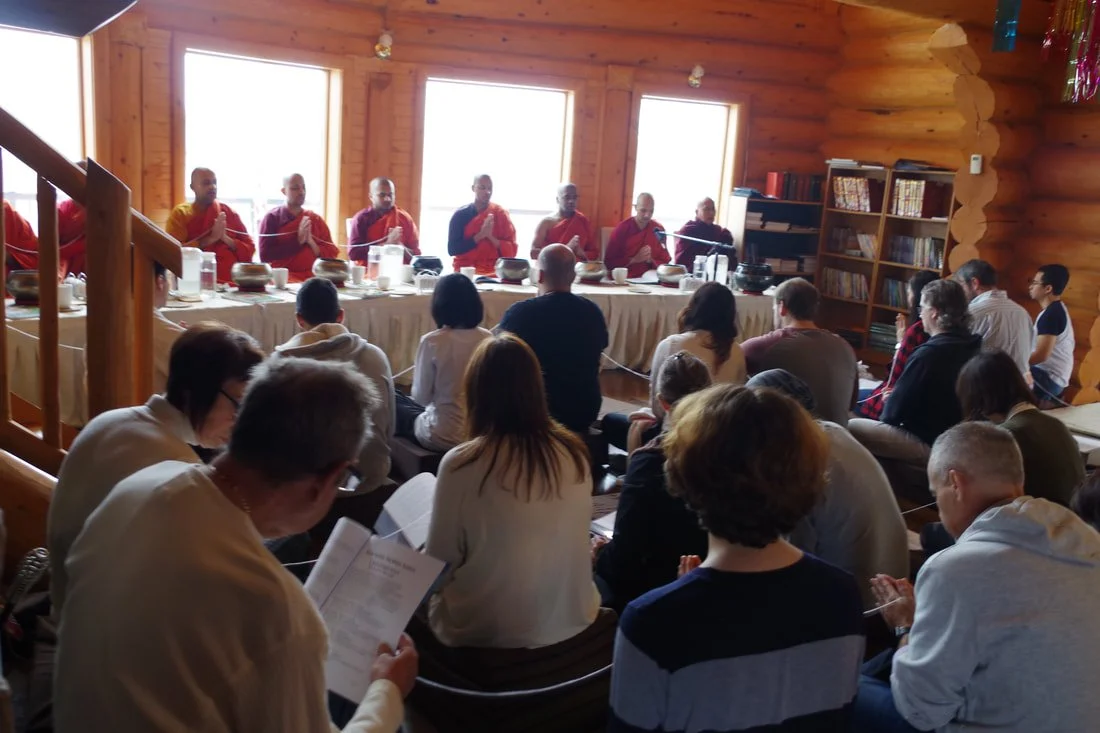 Indoor meditation session with seated monks in red robes and participants in a rustic wooden room, books on shelves in the background.