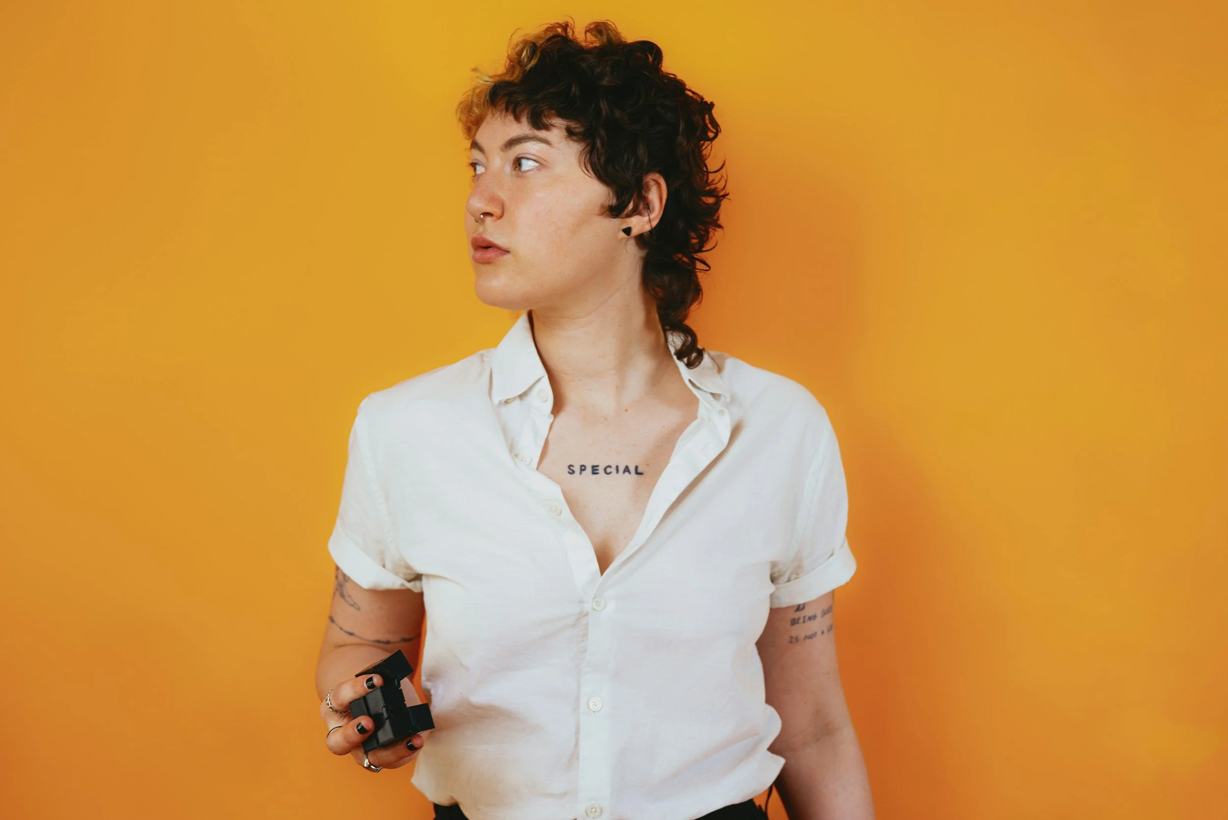 Person with short curly hair, wearing a white shirt, holding a black object, standing against an orange background.