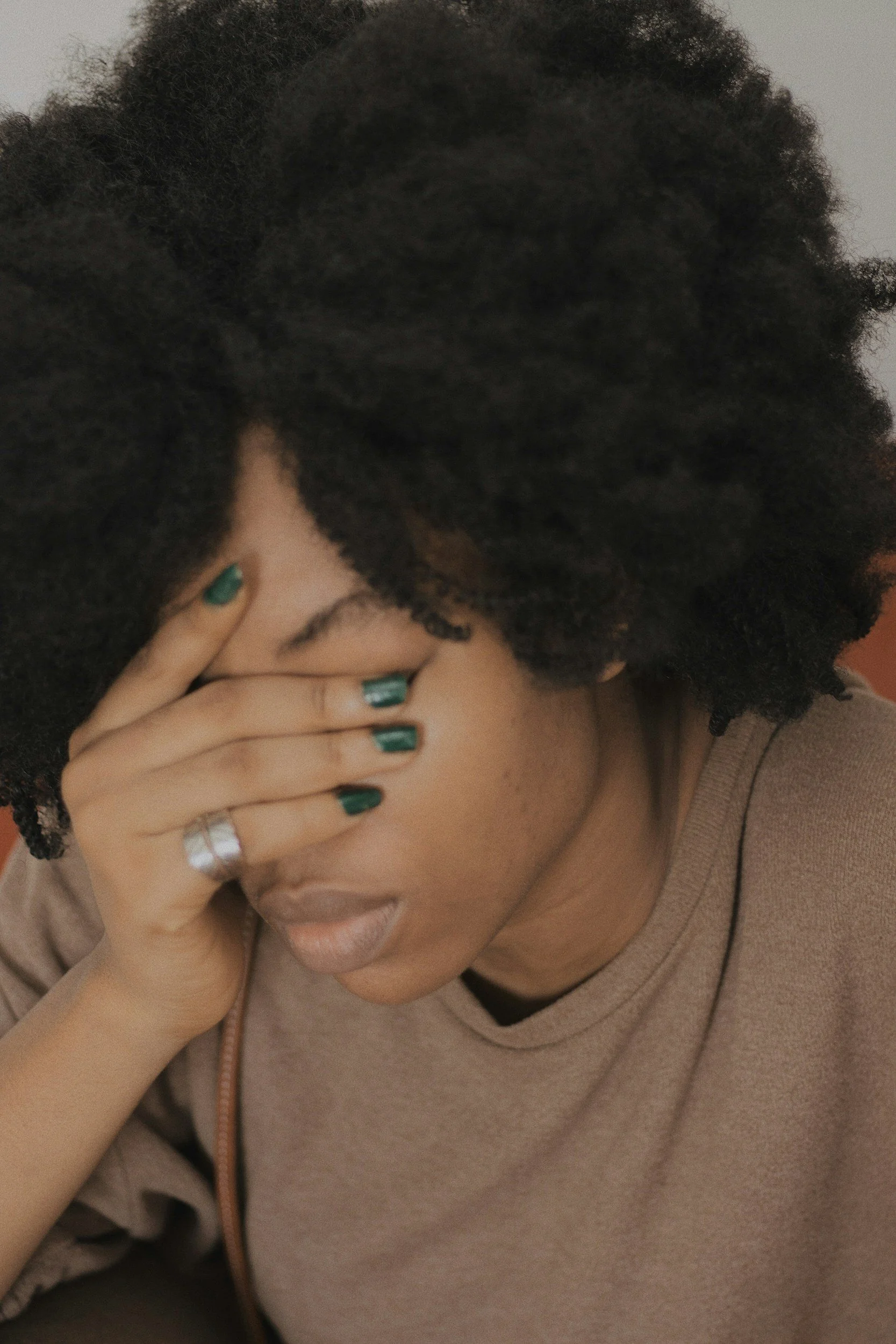A woman with curly black hair covering her face with her hand, wearing a beige top and dark green nail polish.